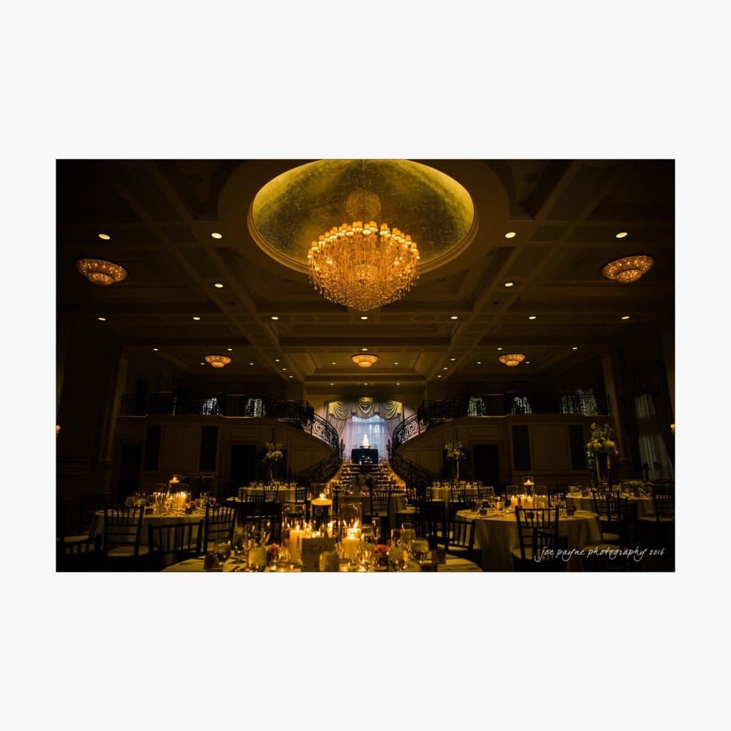 A dimly lit ballroom with elegantly set tables and golden candlelight. In the center, a grand staircase leads to a stage with a backdrop of sheer curtains. A large, ornate chandelier hangs from the ceiling, illuminating the space with a warm glow.