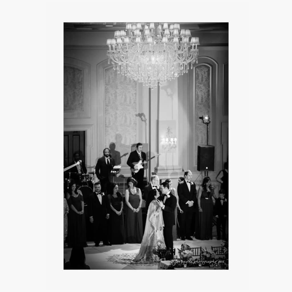 A black-and-white photo of a wedding ceremony in an elegant hall. The couple stands under a large chandelier, surrounded by the wedding party. Musicians are in the background. The atmosphere is formal and celebratory.