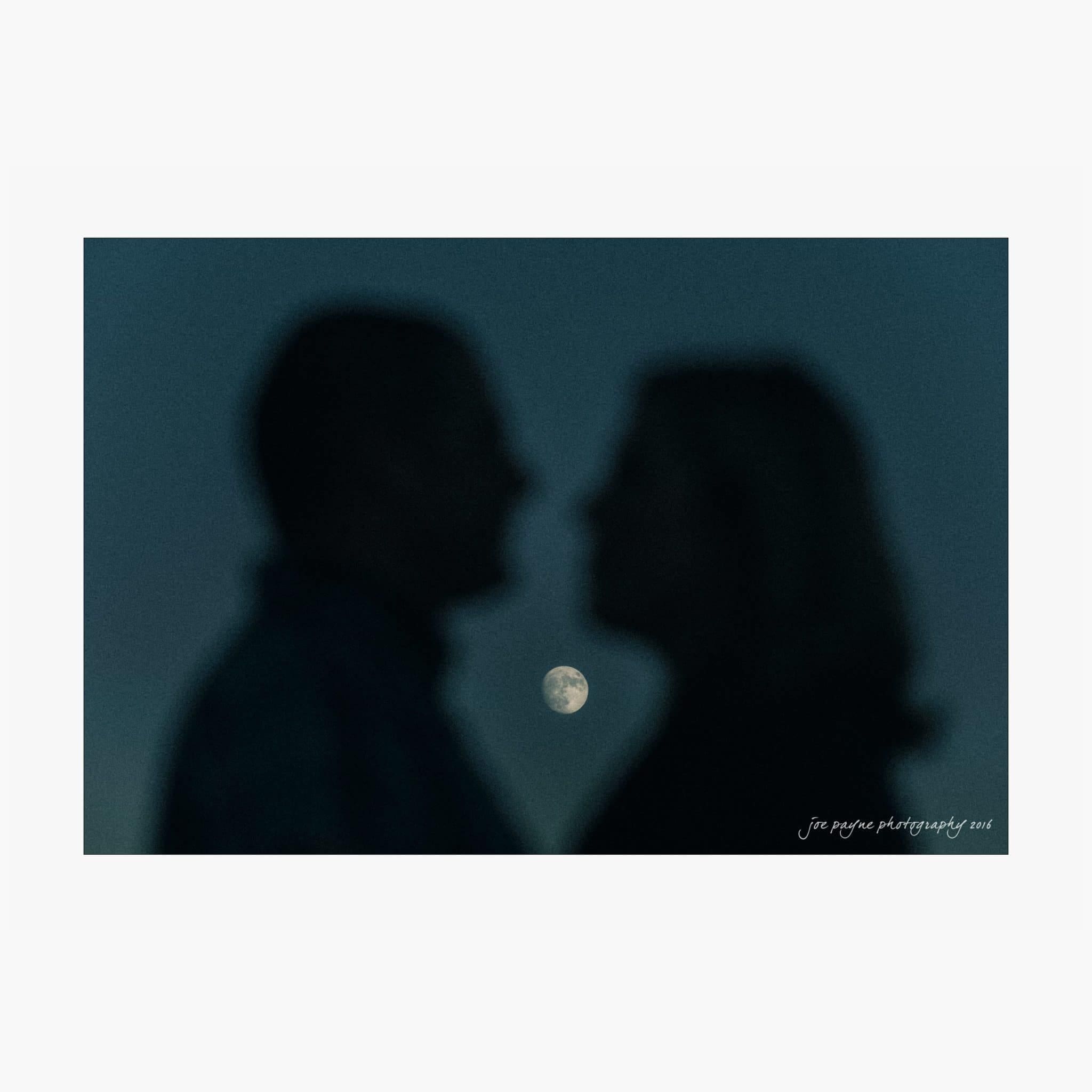 raleigh engagement photography | kim & jeff Silhouettes of two people facing each other in front of a full moon, set against a dark blue sky.