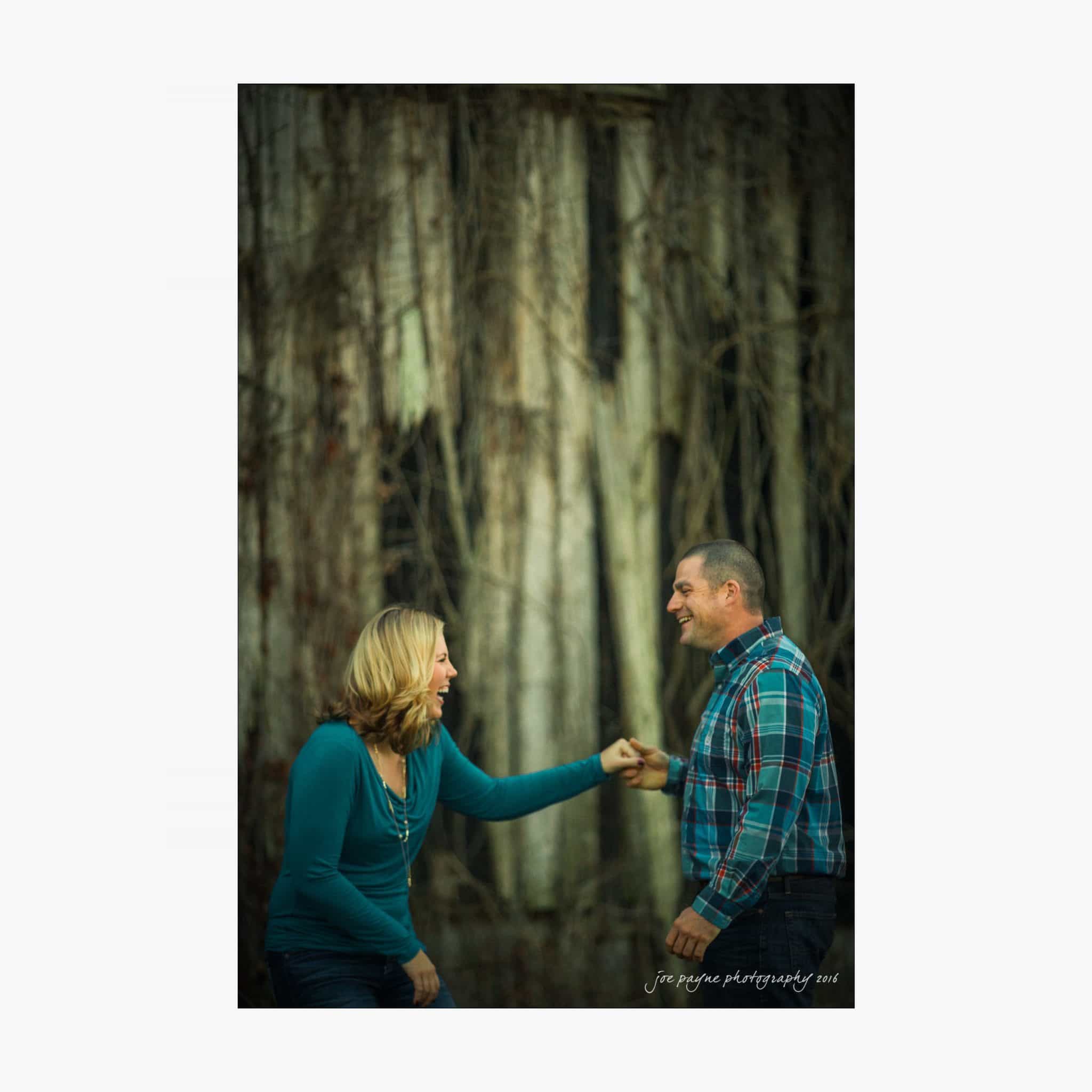 raleigh engagement photography | kim & jeff A woman and a man stand in front of a large, textured tree trunk. The woman, wearing a teal top, gestures animatedly towards the smiling man in a plaid shirt. They appear happy and engaged in conversation.