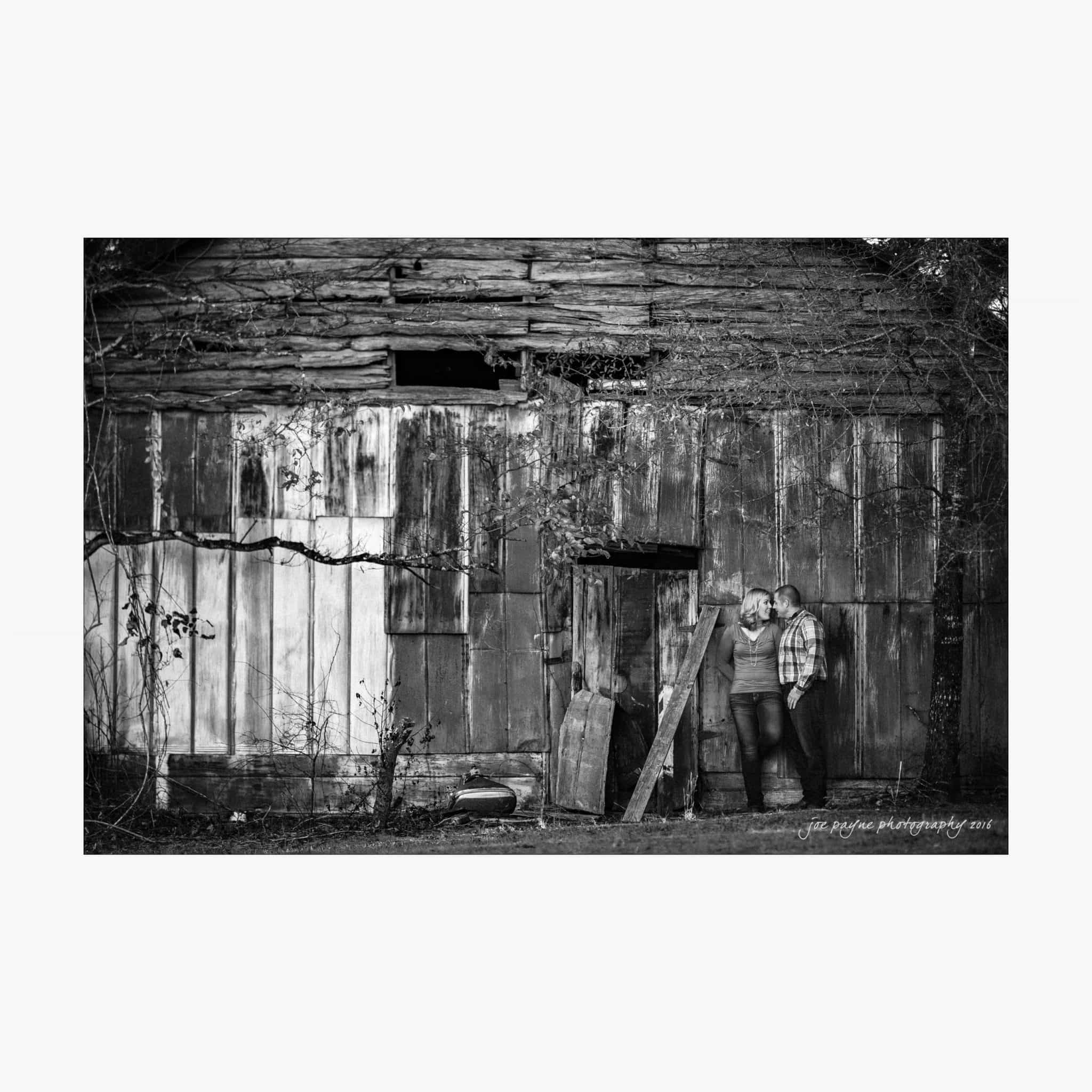 raleigh engagement photography | kim & jeff A couple leans against an old, weathered wooden barn, surrounded by creeping vines. The black-and-white photo captures a rustic and intimate moment, highlighting the texture of the barns planks and the couples casual pose.