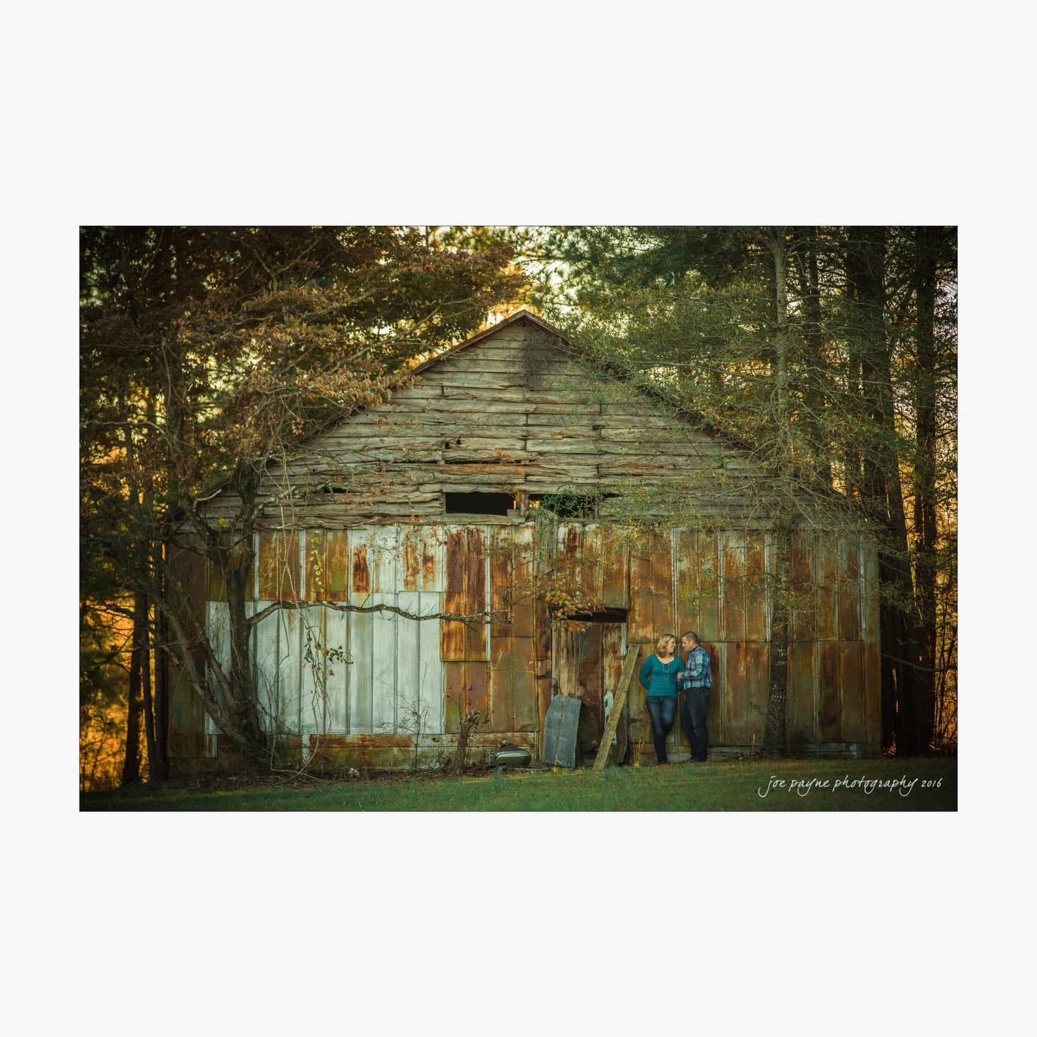raleigh engagement photography | kim & jeff A couple stands close together in front of an old, weathered barn. The barns rusted exterior is surrounded by trees. The setting sun casts a warm glow on the scene, creating a peaceful, rustic atmosphere.