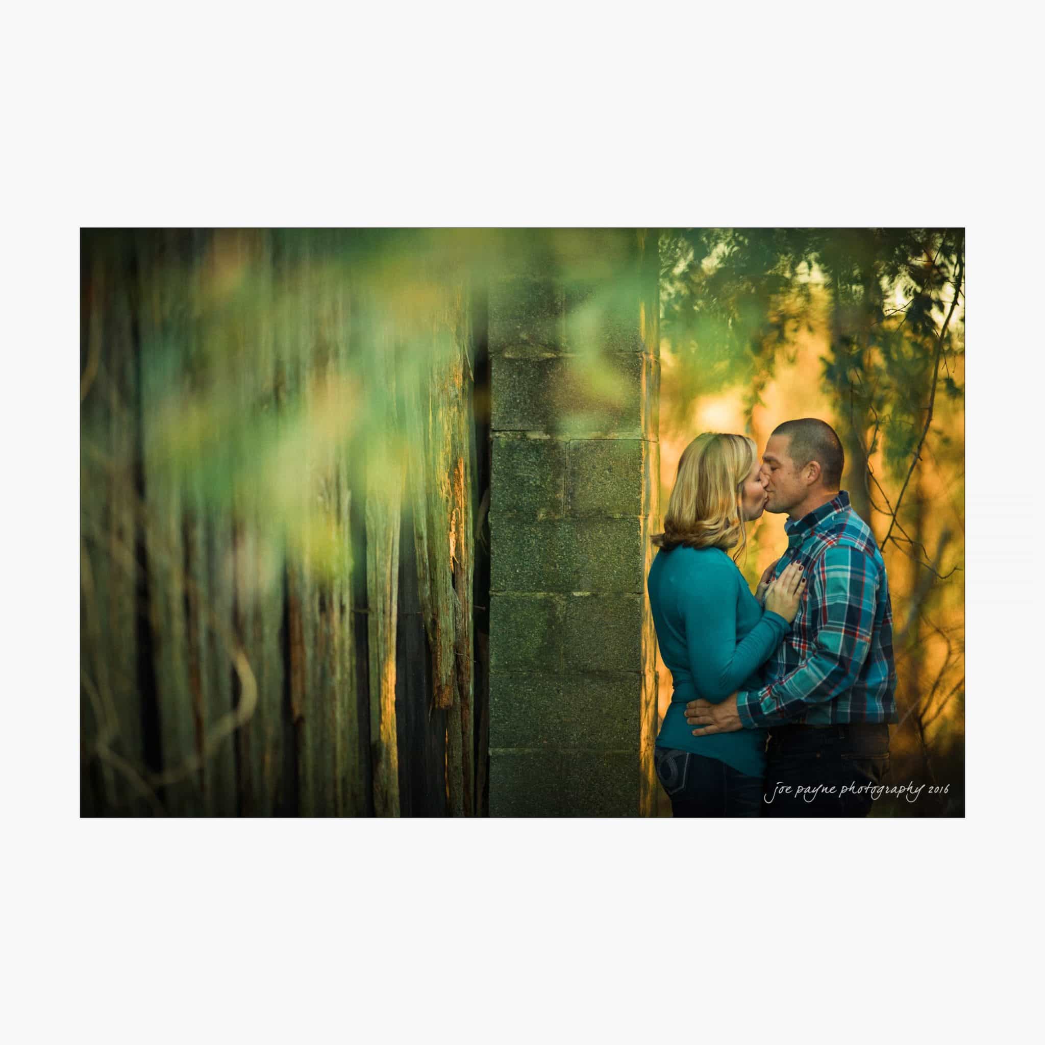 raleigh engagement photography | kim & jeff A couple embraces near an old wooden wall, surrounded by blurred greenery. They stand close, gazing into each others eyes under soft, warm light. The man wears a plaid shirt, and the woman is in a blue sweater. The scene is intimate and peaceful.