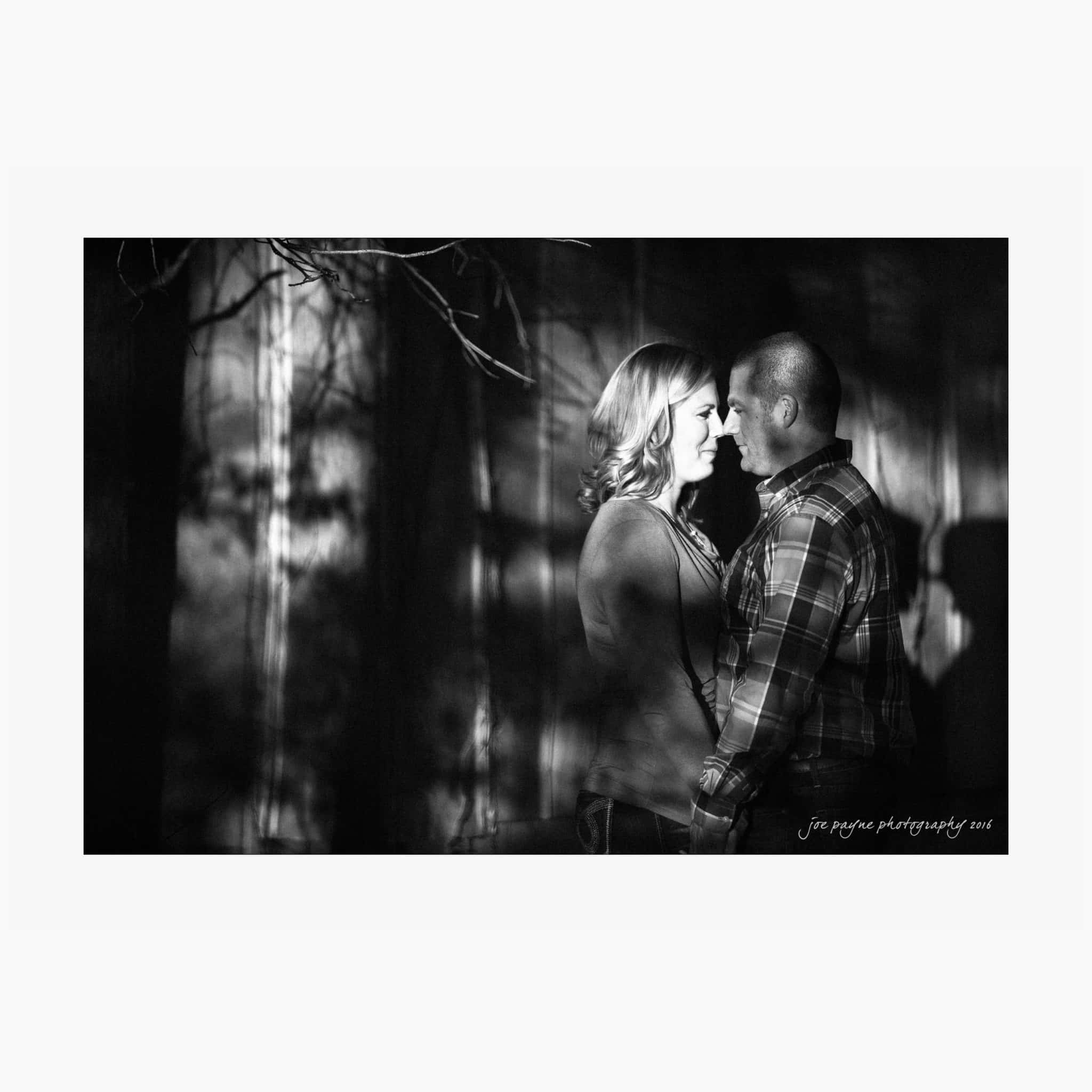 raleigh engagement photography | kim & jeff A couple stands facing each other in a black and white photo, illuminated by dappled sunlight. The background is a blurred natural setting, creating a serene and intimate atmosphere.