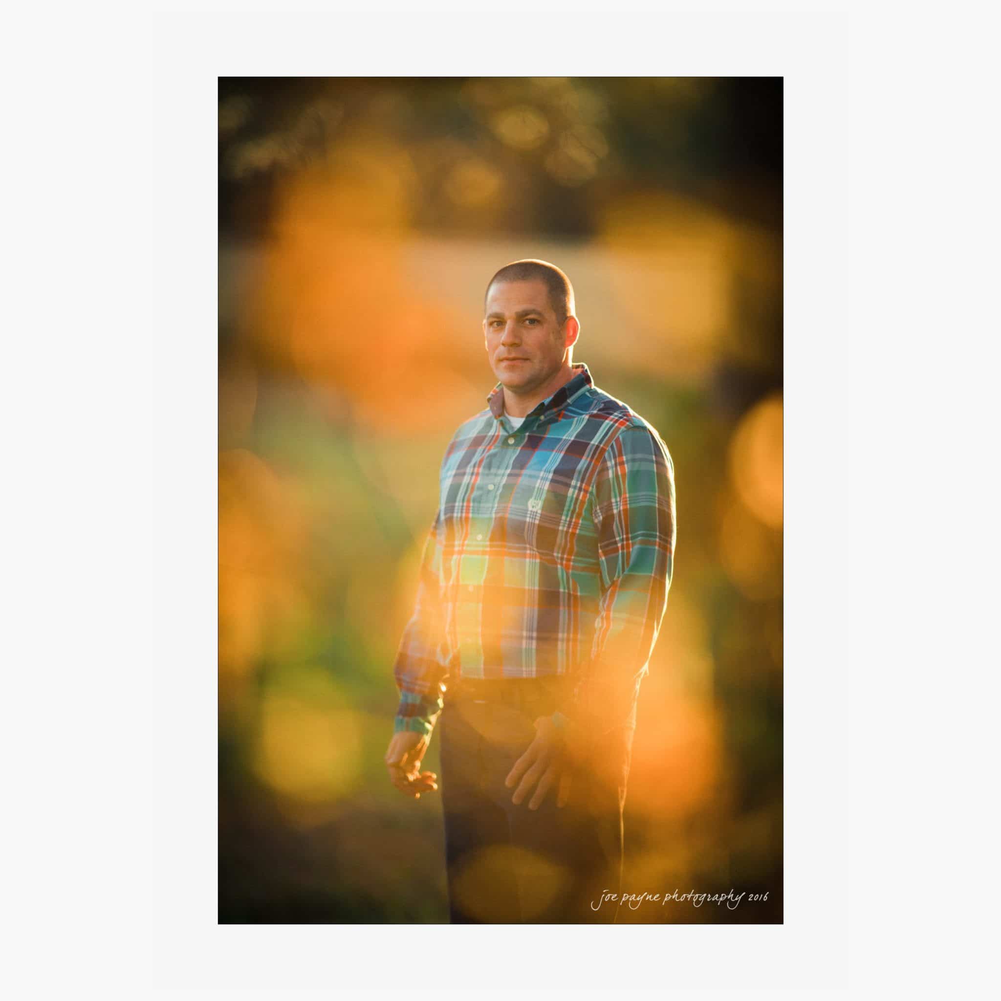 raleigh engagement photography | kim & jeff A person wearing a plaid shirt stands outdoors with a serene expression. The image has a warm, golden bokeh effect that adds a soft, sunny glow to the scene.