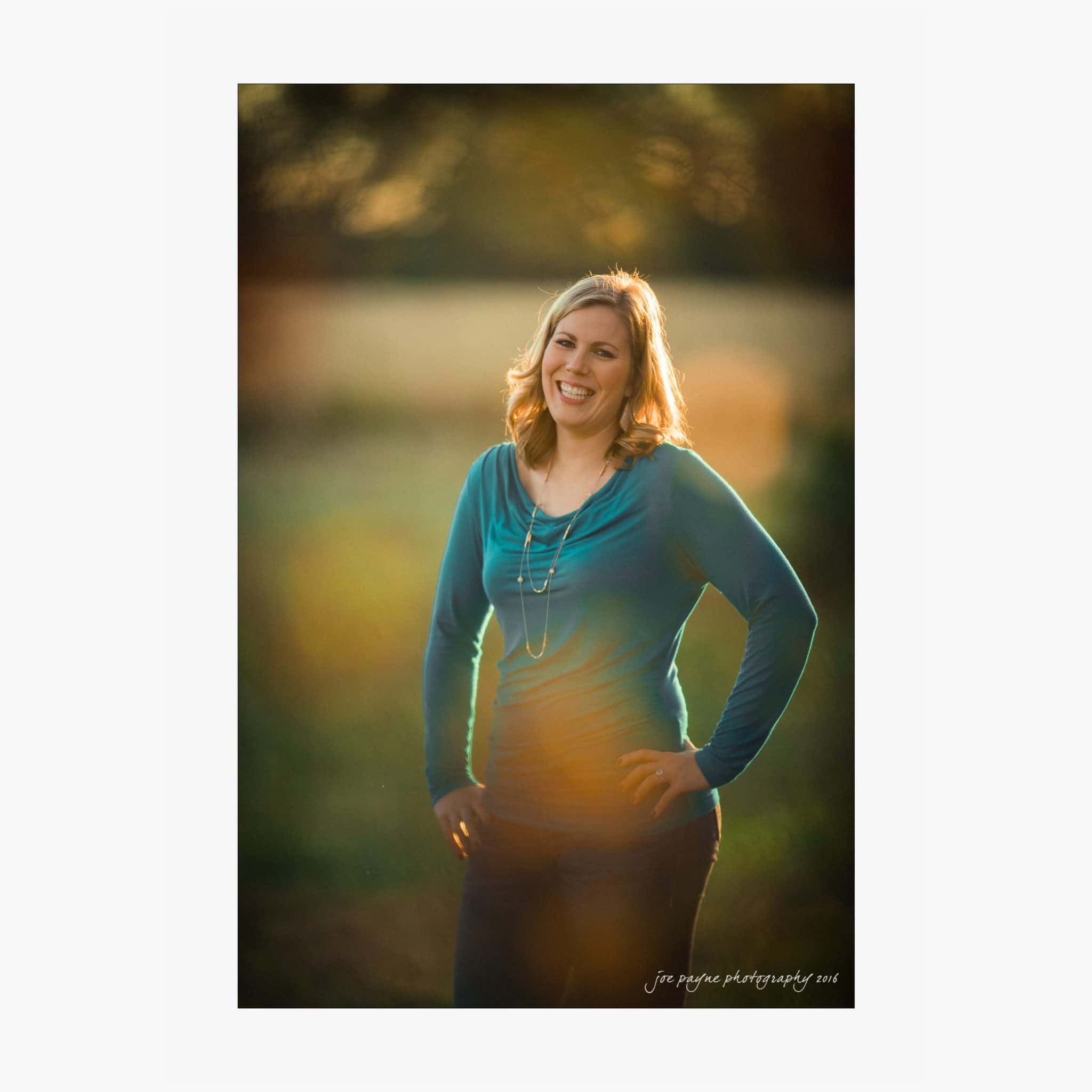 raleigh engagement photography | kim & jeff A woman with shoulder-length blonde hair stands outdoors, smiling, with hands on hips. She is wearing a blue long-sleeved top and a necklace. The background is softly blurred greenery.