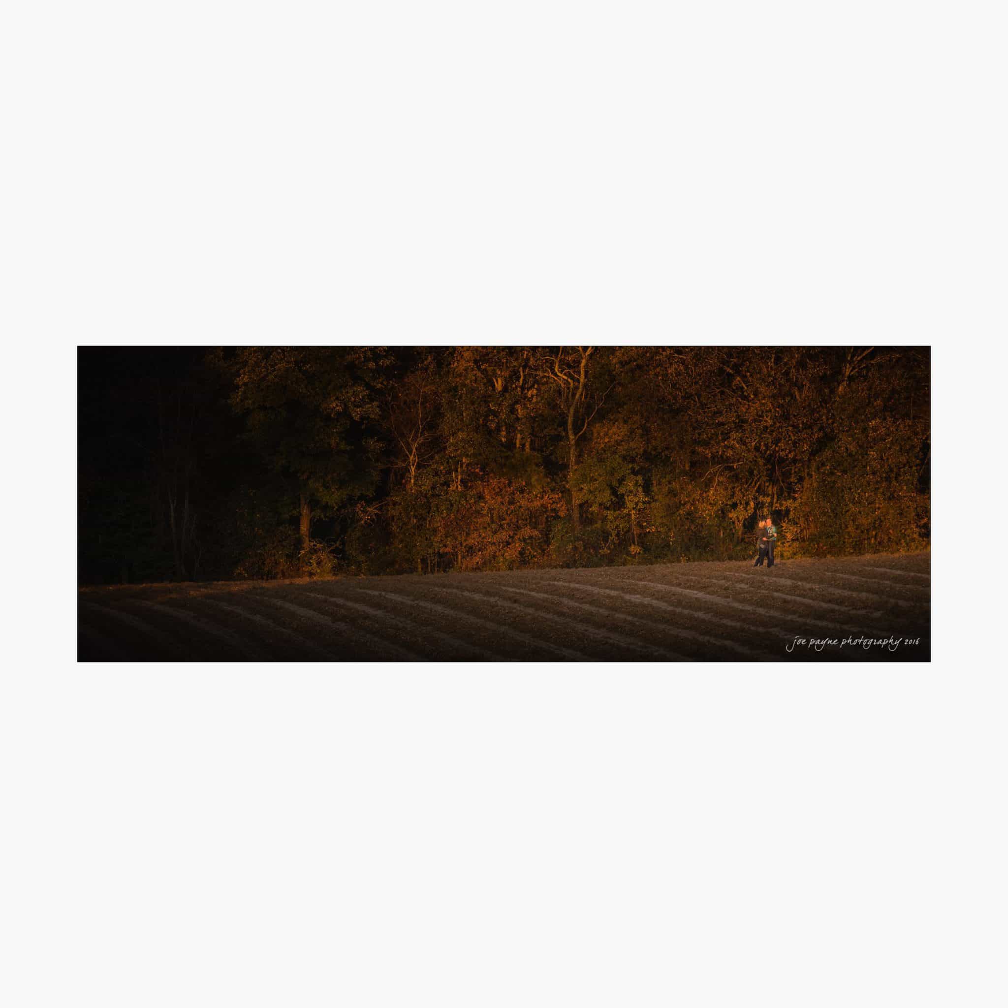 raleigh engagement photography | kim & jeff A person walks along the edge of a field at sunset, surrounded by trees with autumn foliage. The warm glow of the setting sun casts a golden hue over the landscape, highlighting the textures of the field and trees.