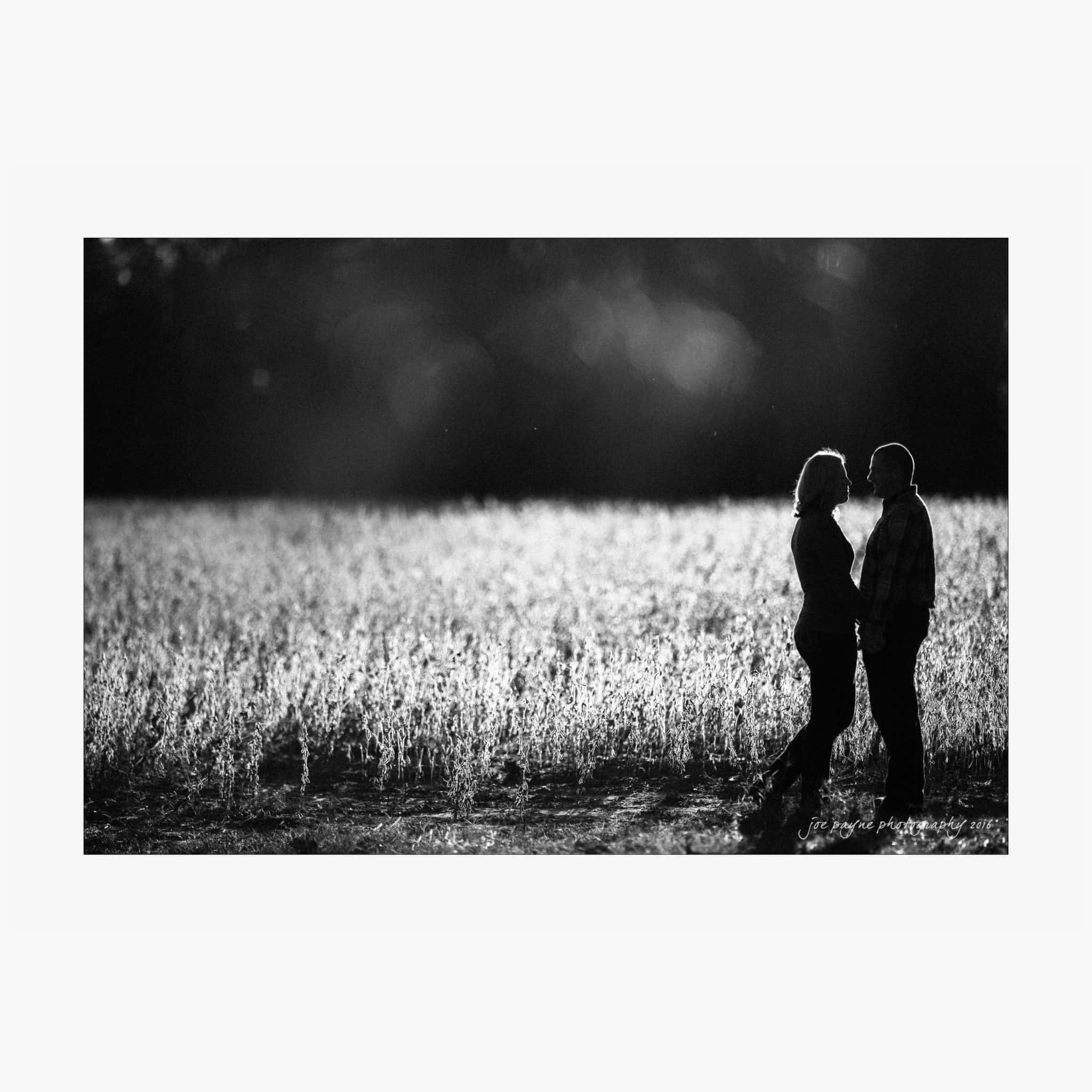 raleigh engagement photography | kim & jeff A black and white image of a couple standing in a sunlit field facing each other. The field is surrounded by a dark, blurred background, highlighting the light on the grass and the silhouettes of the couple.