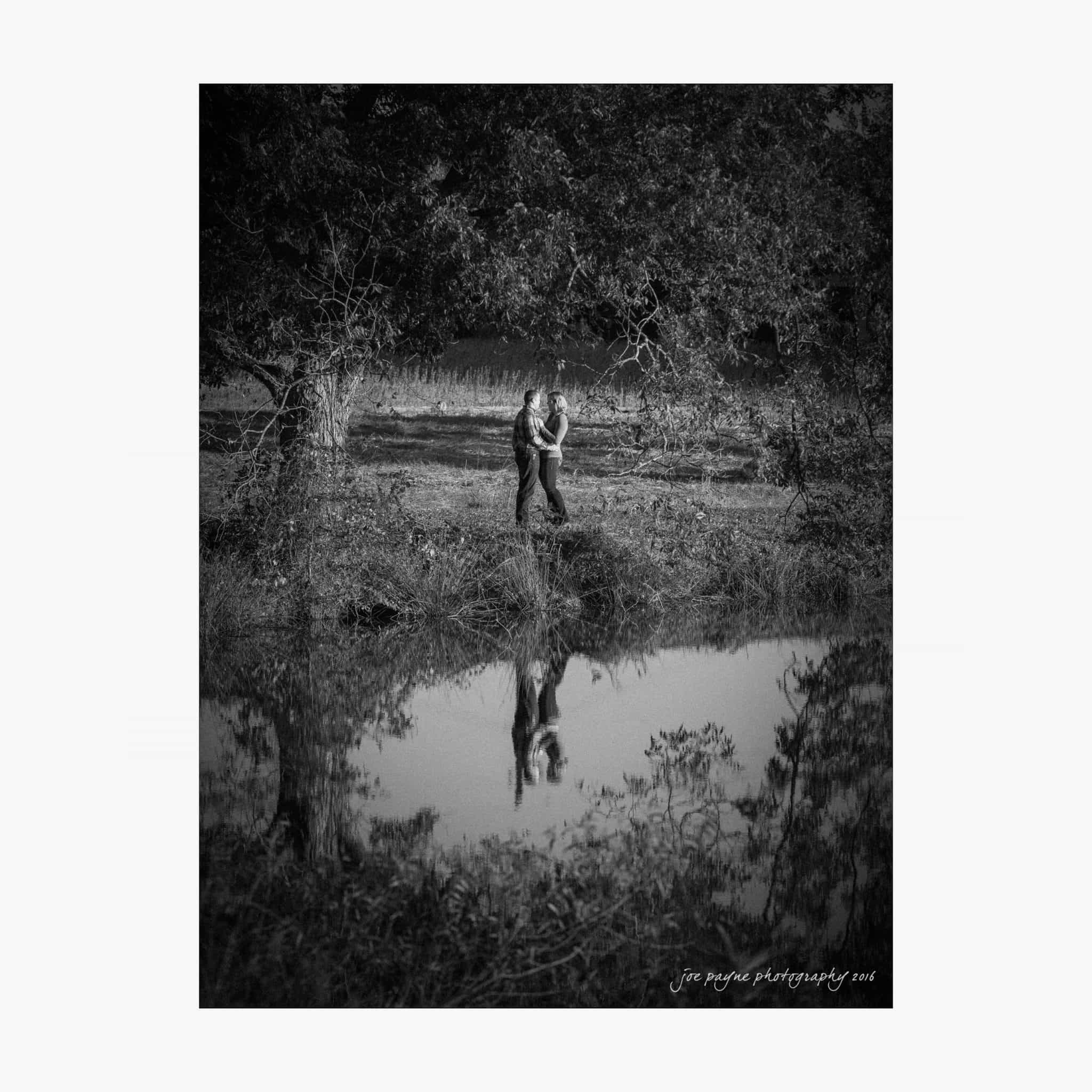raleigh engagement photography | kim & jeff A black and white photo of a couple embracing near a tree by a pond. The couples reflection is visible in the water, with lush foliage around them, creating a serene and romantic atmosphere.