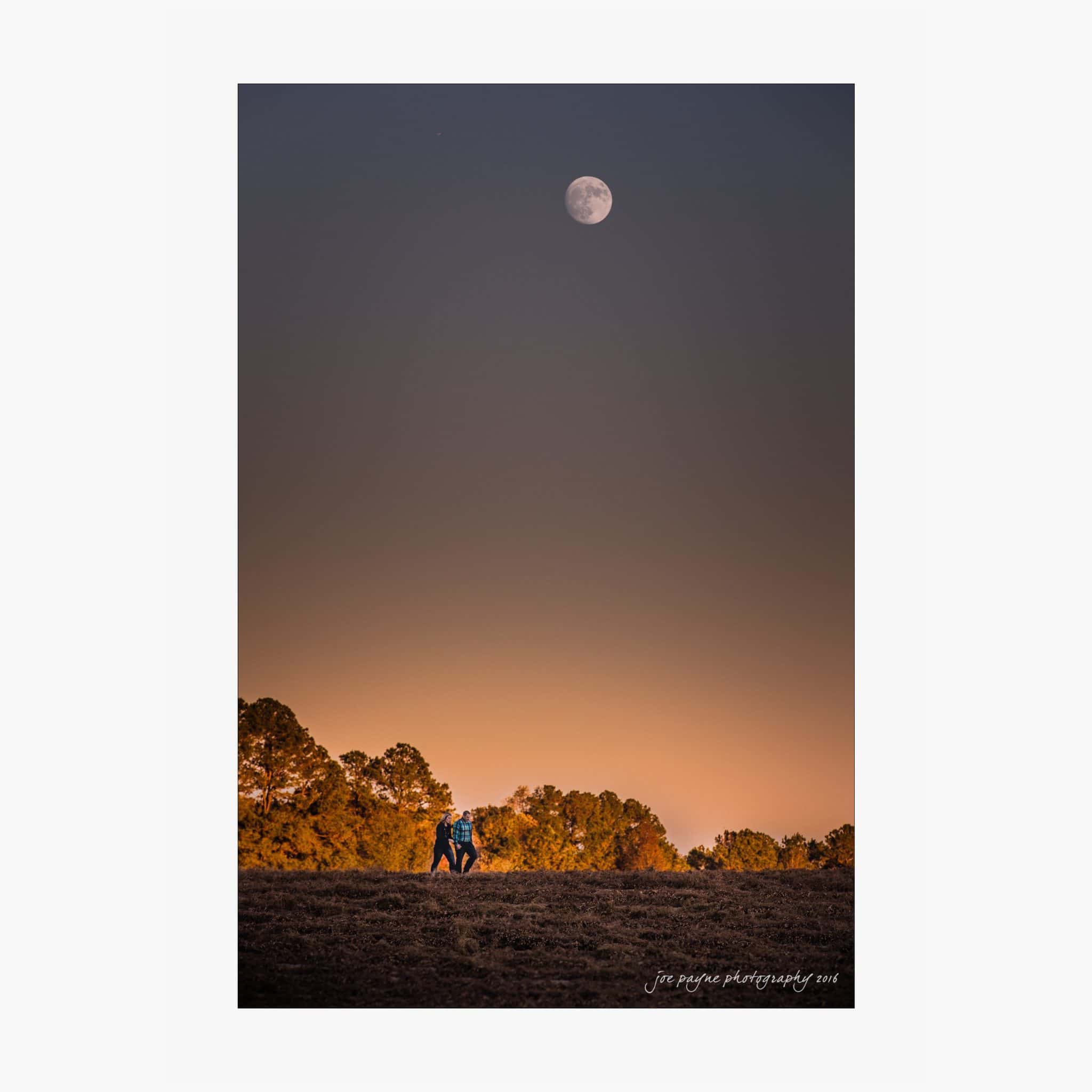 raleigh engagement photography | kim & jeff A couple walks hand in hand across a field at dusk, with trees lining the background. The sky above is deep orange and purple, featuring a bright full moon.