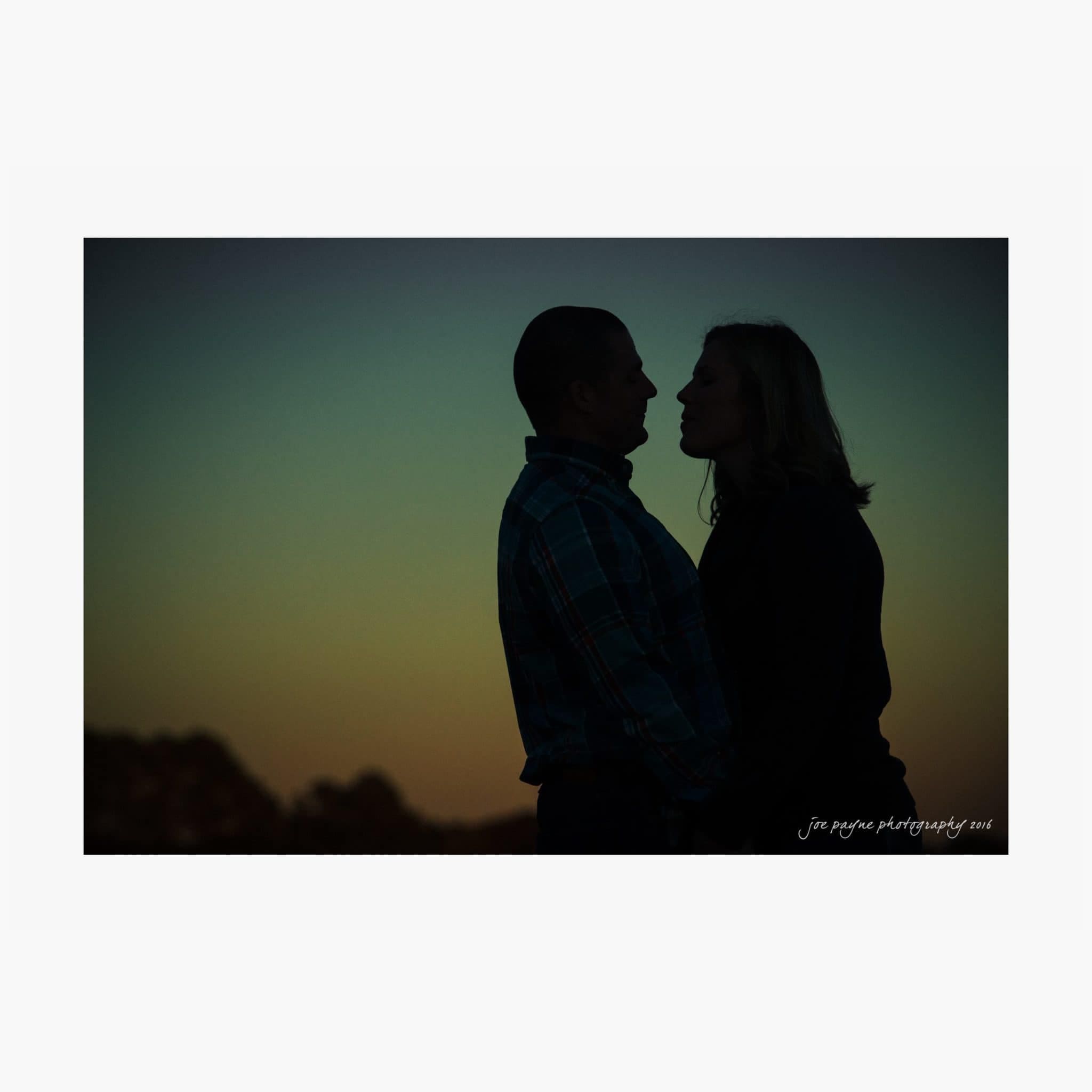 raleigh engagement photography | kim & jeff Silhouetted couple standing close, facing each other against a twilight sky with a gradient of blue and orange hues.