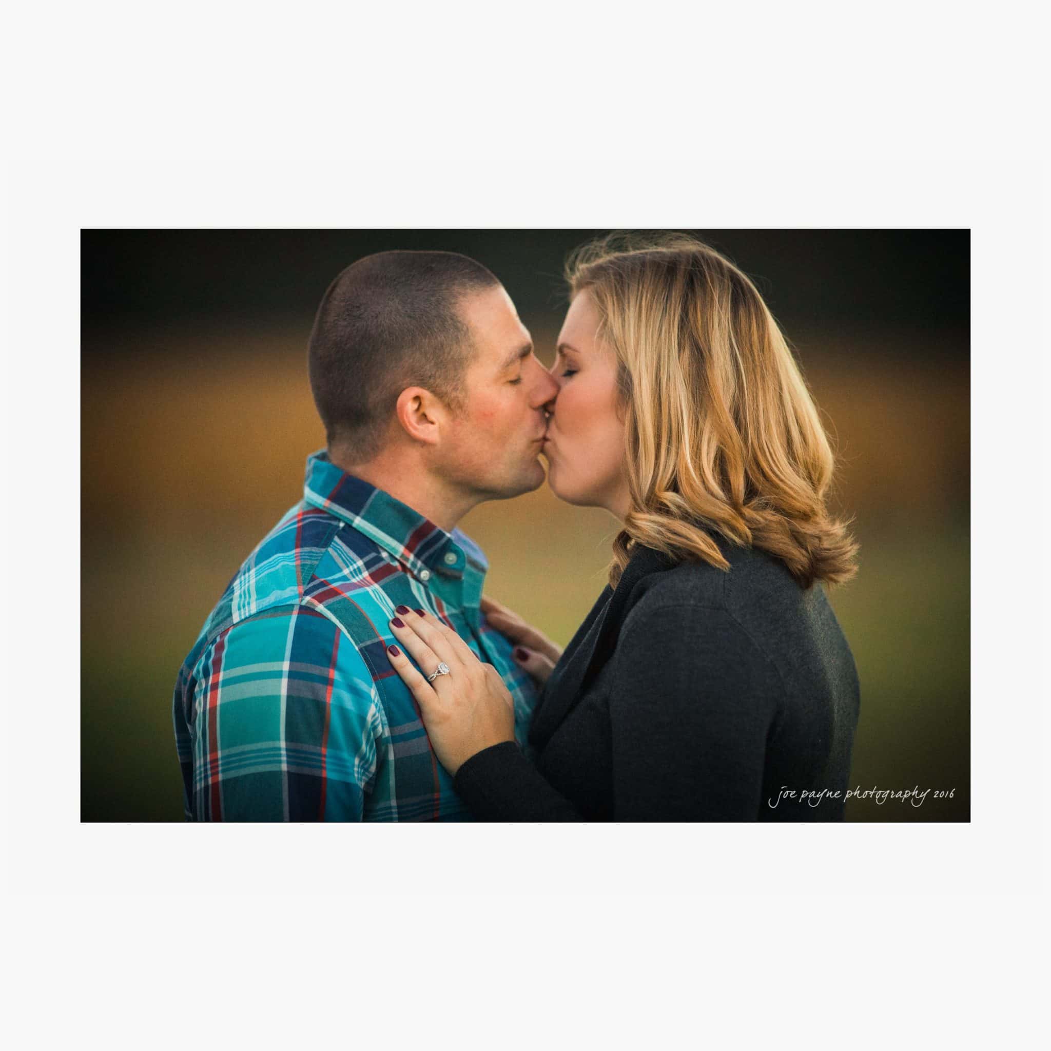 raleigh engagement photography | kim & jeff A couple shares a kiss outdoors. The man wears a plaid shirt and has short hair. The woman has shoulder-length blonde hair and wears a dark jacket. She has an engagement ring on her left hand, which rests on his shoulder. The background is blurred.