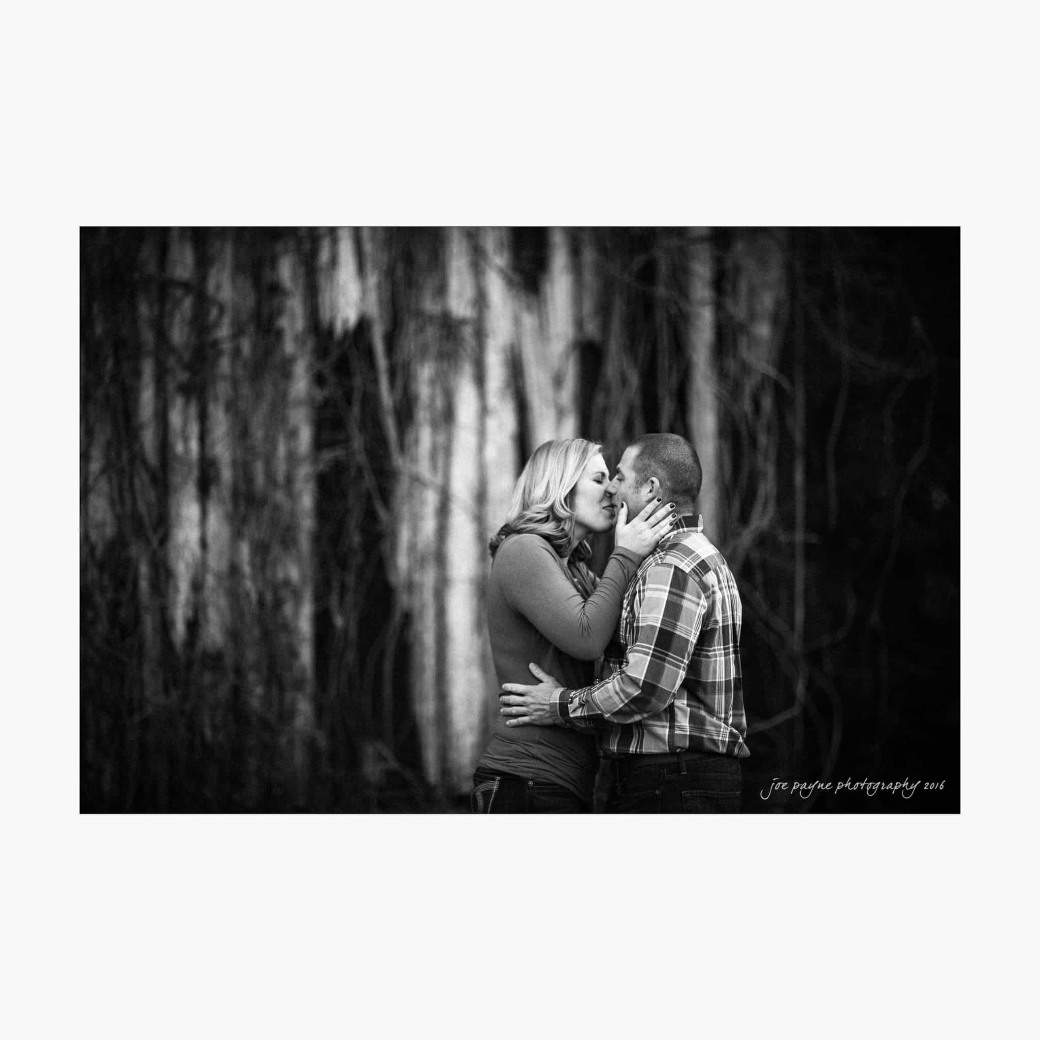 raleigh engagement photography | kim & jeff A couple stands close, sharing a tender moment in a wooded area. The woman gently holds the mans face as they embrace. The background features blurred trees, creating a romantic atmosphere in this black and white photograph.
