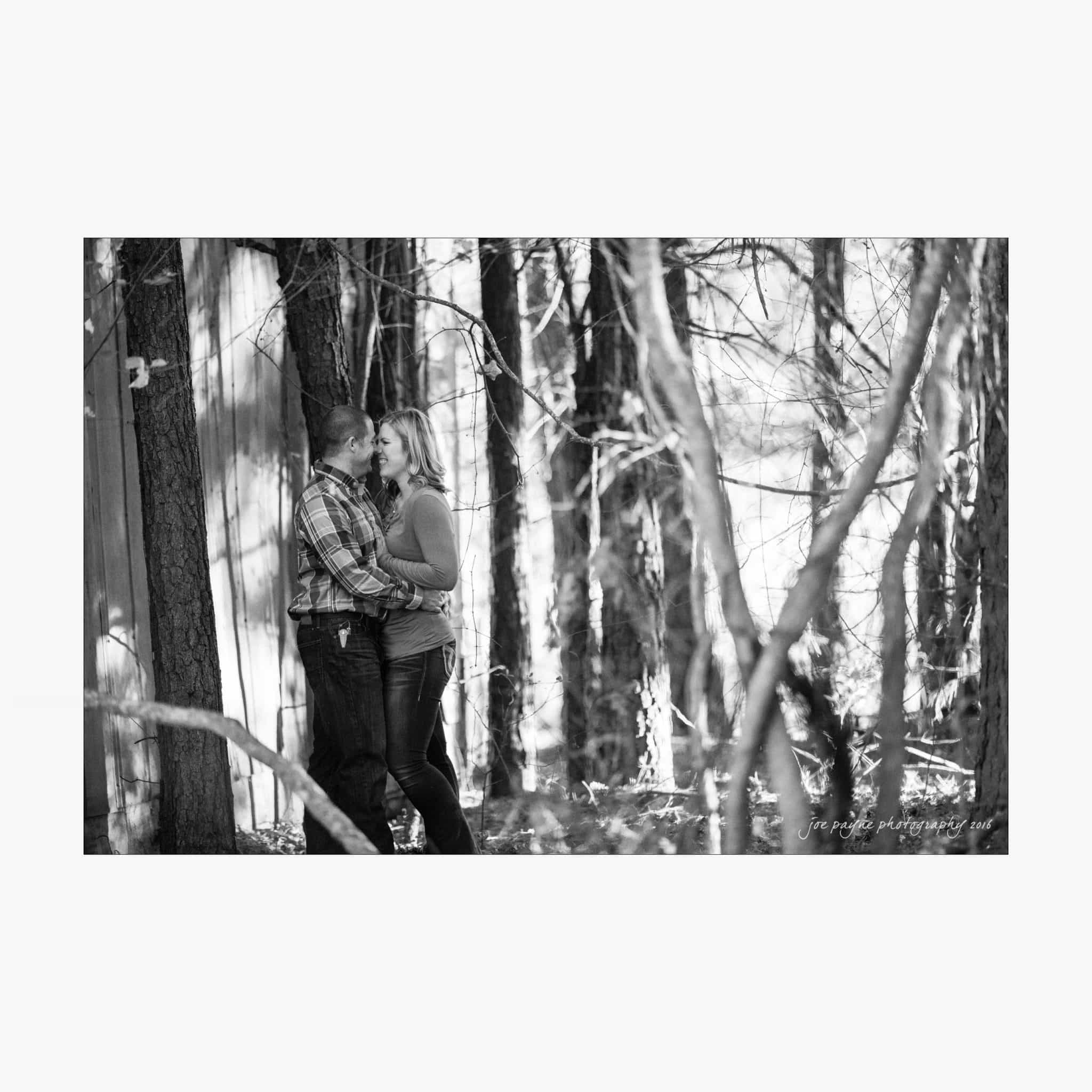 raleigh engagement photography | kim & jeff A couple stands in a forest, leaning against a tree and kissing. Sunlight filters through the trees, creating a serene and intimate atmosphere. The image is in black and white.
