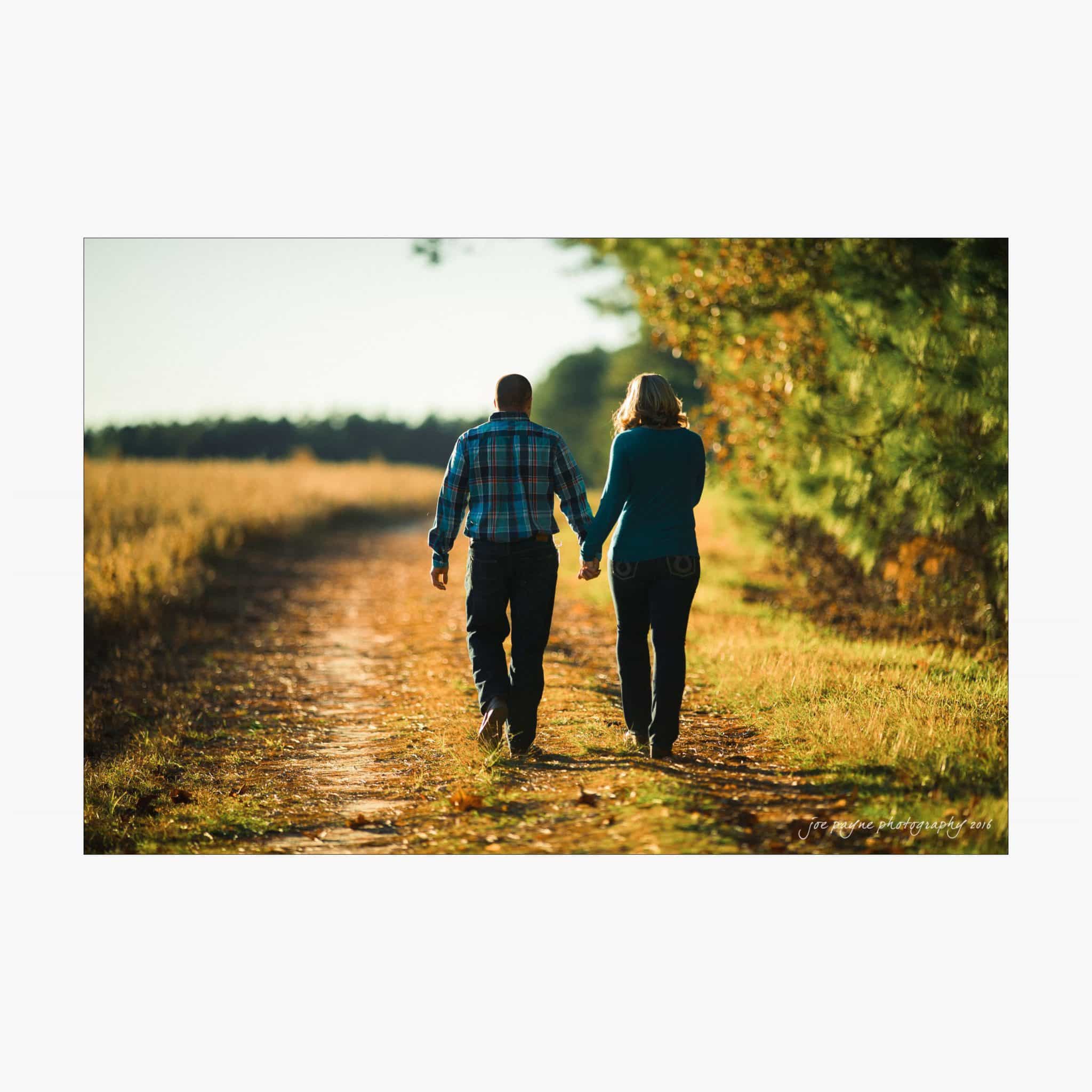 raleigh engagement photography | kim & jeff A couple holding hands while walking down a rustic path flanked by tall grass on one side and dense trees on the other. They are both wearing casual clothes, and the sunlight casts long shadows on the path.