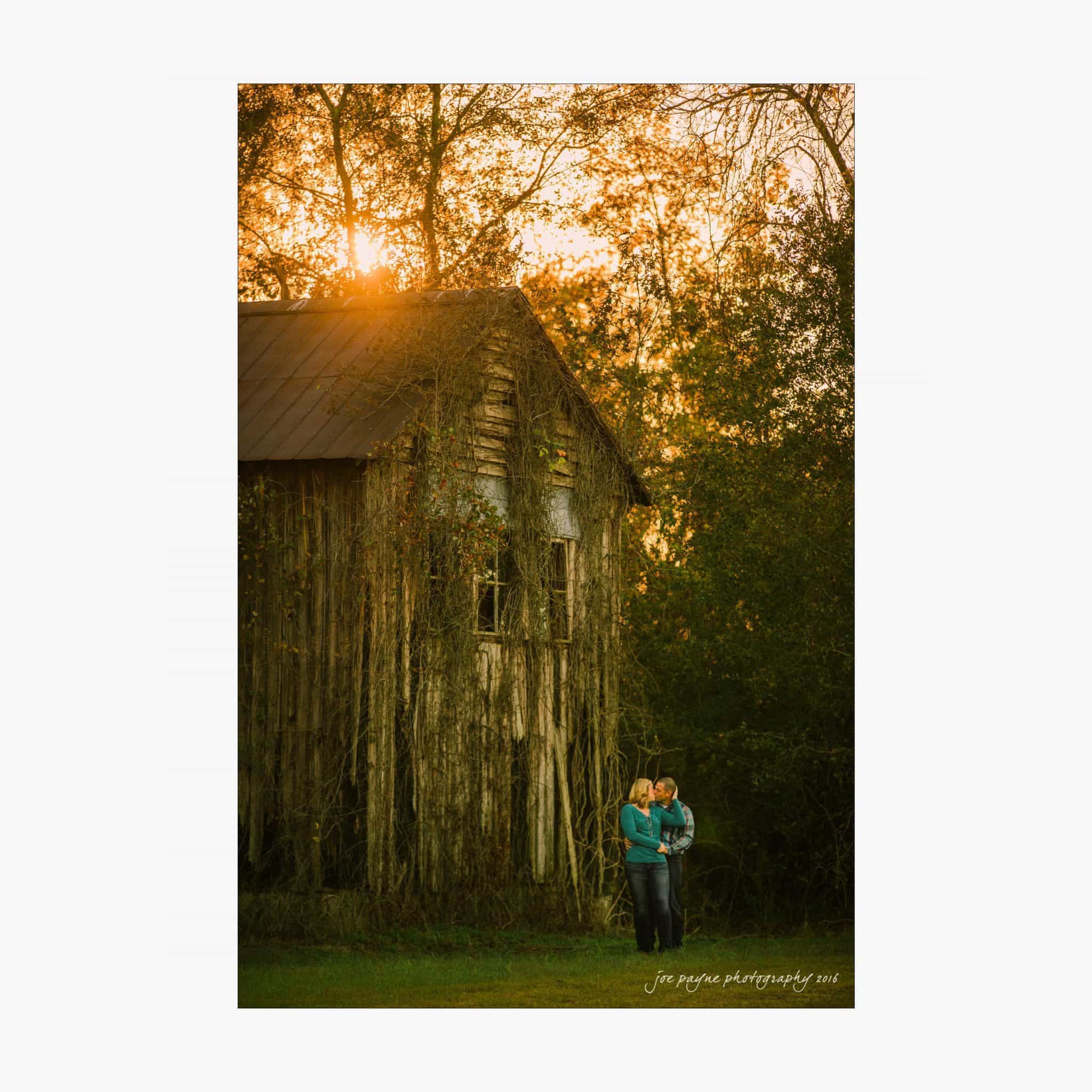 raleigh engagement photography | kim & jeff A couple embraces in front of an old barn covered in vines, with the warm glow of the setting sun shining through the trees. The scene exudes a peaceful, rustic charm.