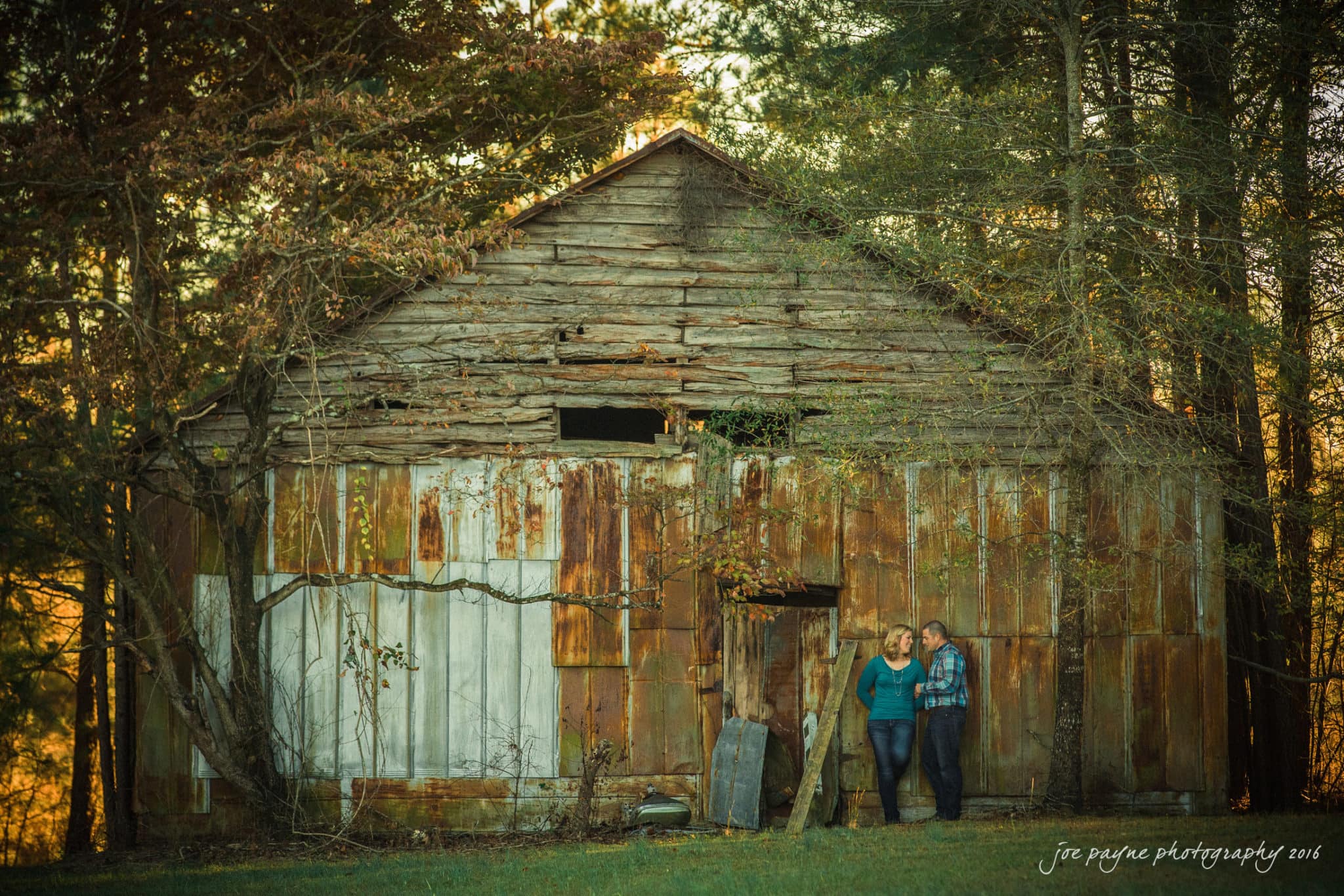 A couple stands side by side in front of an old, weathered barn, surrounded by tall trees and overgrown plants in the warm, golden light of late afternoon—a perfect scene for Raleigh engagement photography.
