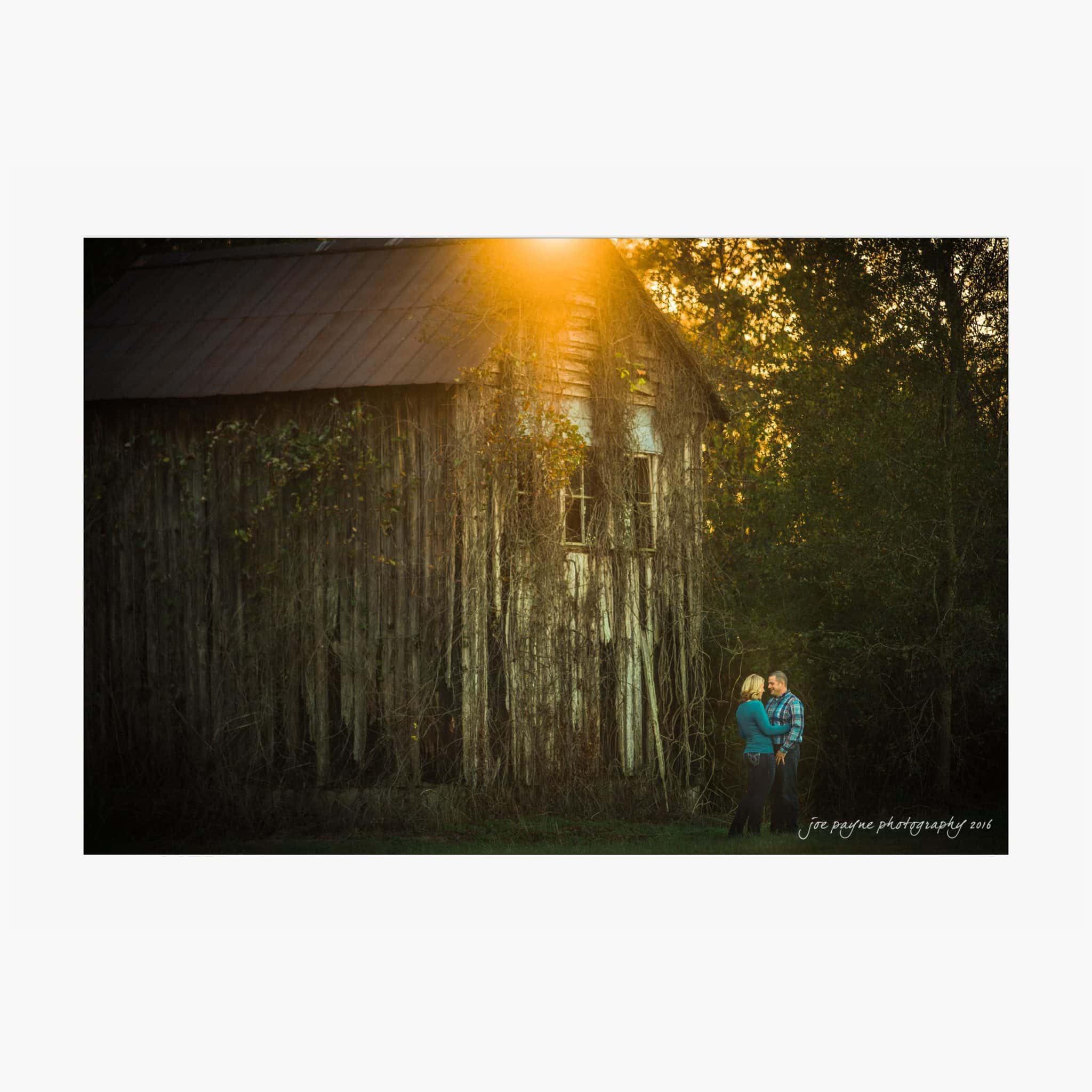raleigh engagement photography | kim & jeff A couple stands close together in front of an old, ivy-covered wooden barn. The sun sets in the background, casting a warm glow over the scene. The surrounding trees enhance the rustic, intimate atmosphere.