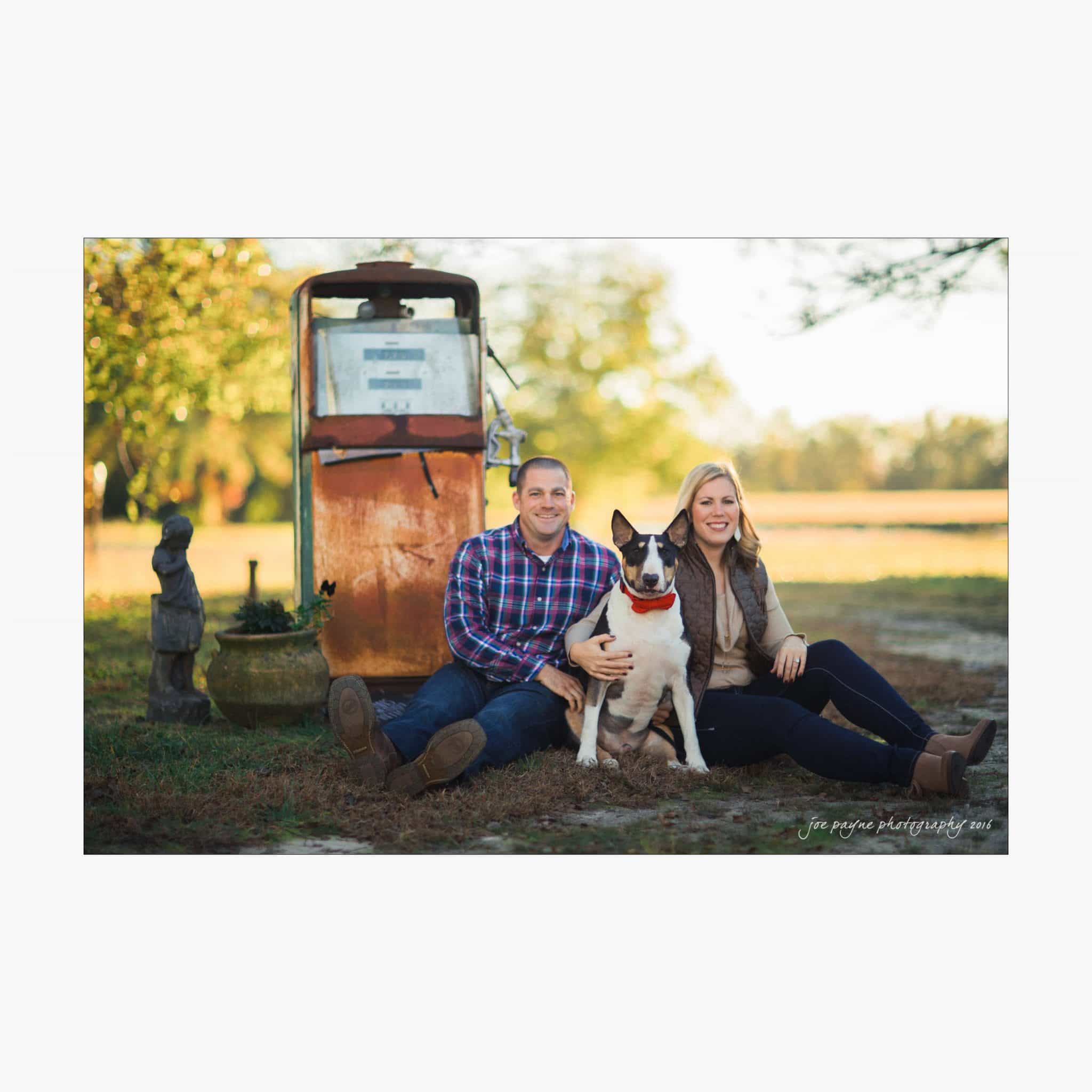 raleigh engagement photography | kim & jeff A couple sitting on grass with a black and white dog between them, beside an old gas pump. They are outdoors with trees in the background, and both are smiling. The man wears a plaid shirt and jeans, and the woman wears a jacket and scarf.