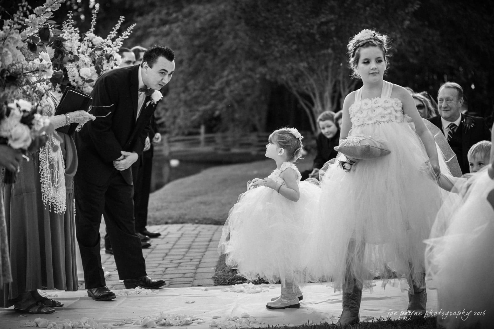 shady wagon farm wedding photography - danielle & josh A black and white photo of a wedding ceremony. Two young girls in fluffy dresses walk down an aisle. The man in a suit bends down, smiling. Guests seated on both sides watch. Lush greenery and floral decorations surround the scene.