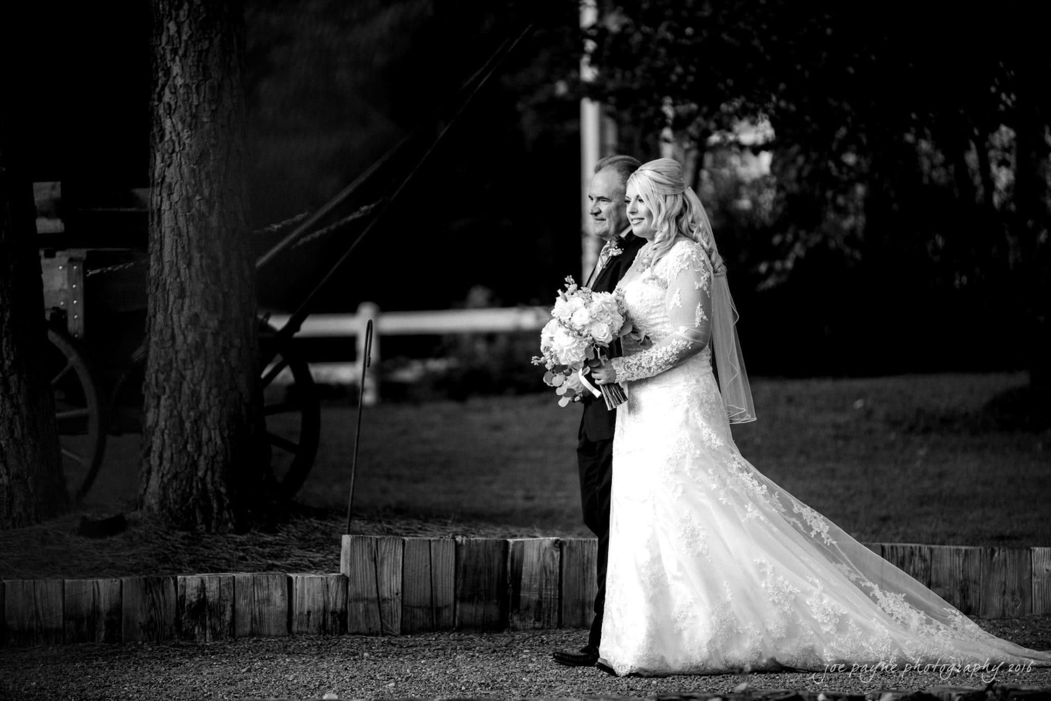 shady wagon farm wedding photography - danielle & josh A bride in a lace gown and veil holds a bouquet as she walks with an older man in a suit. They are outdoors, surrounded by trees, with a vintage carriage visible in the background. The image is in black and white.