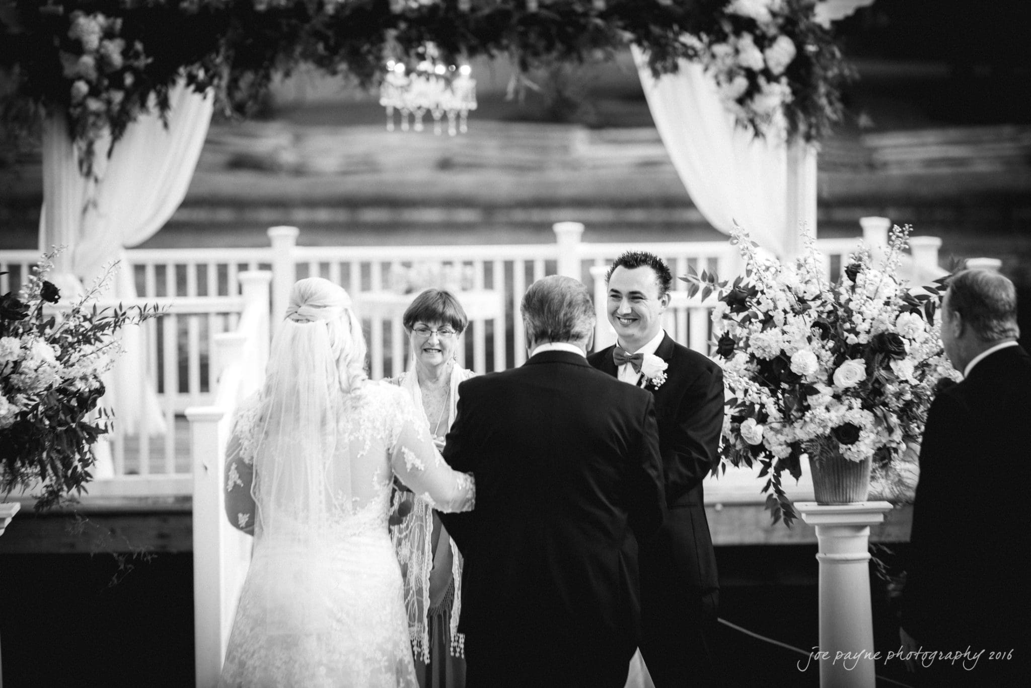 shady wagon farm wedding photography - danielle & josh A black and white photograph of a wedding ceremony. A bride in a gown with a veil stands with her father, facing the groom and officiant. Floral arrangements and draped curtains decorate the outdoor setting. The scene is joyful and elegant.