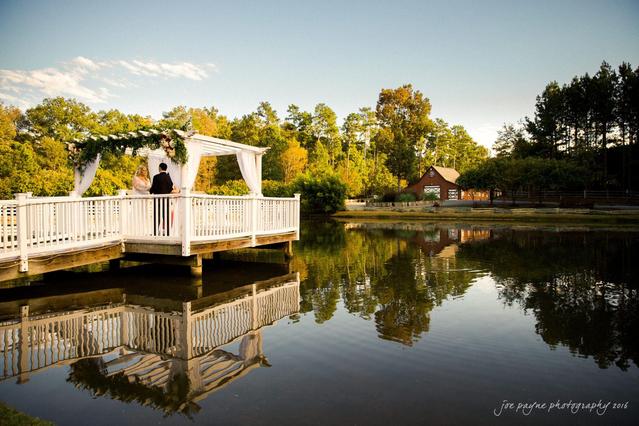 shady wagon farm wedding photography - danielle & josh A couple stands on a white gazebo decorated with flowers by a serene lake, surrounded by trees with autumn foliage. A small building and wooden fence are visible in the background under a clear blue sky.