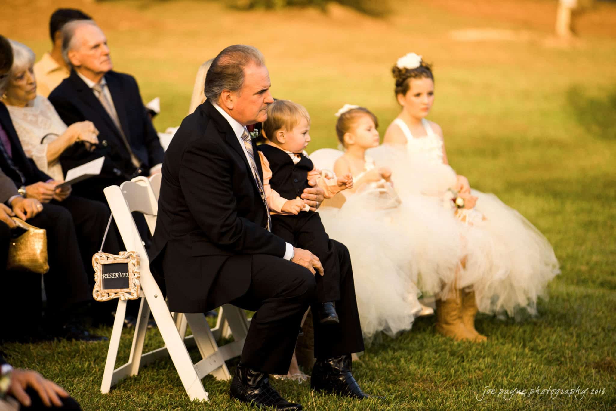shady wagon farm wedding photography - danielle & josh A man in a suit sits on a white chair holding a toddler in formal attire at an outdoor event. Two young girls in white dresses and flowers in their hair sit nearby. Other seated guests are visible in the background.