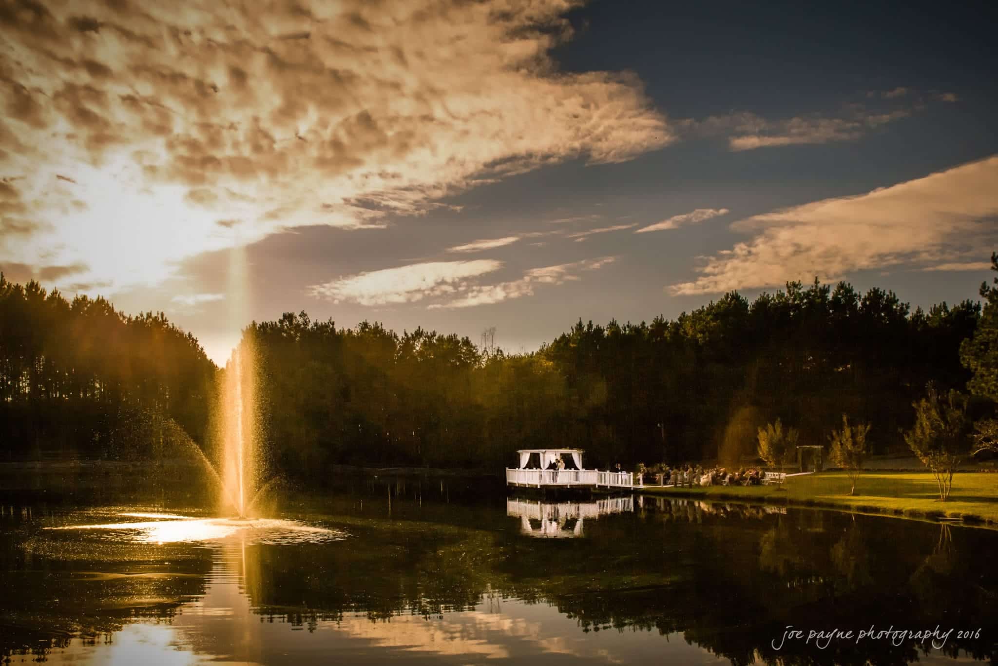 shady wagon farm wedding photography - danielle & josh A serene landscape at sunset featuring a fountain in a tranquil lake. The sky is filled with dramatic clouds. A white gazebo with people gathered nearby is partially reflected in the water, all surrounded by a line of trees.