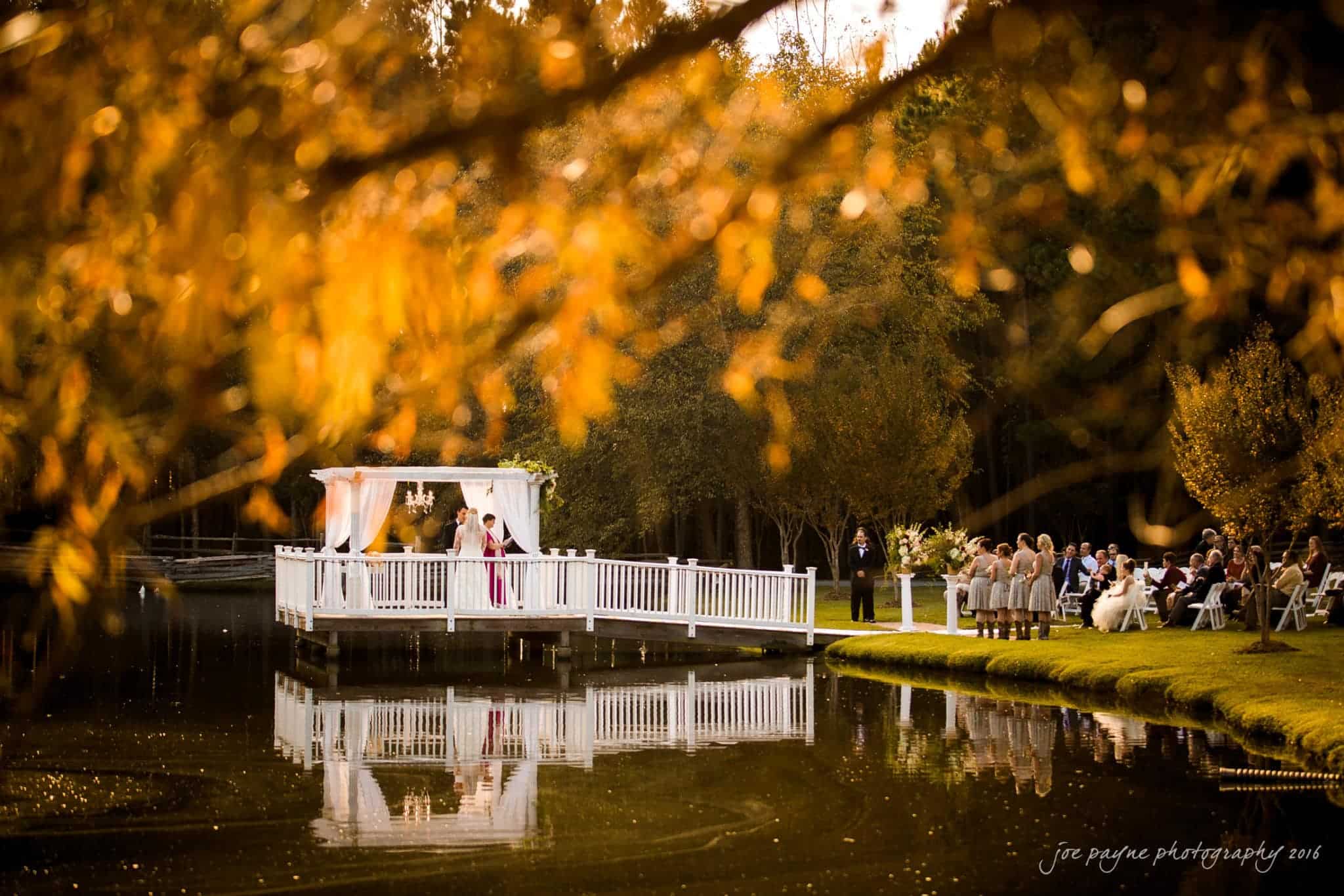 shady wagon farm wedding photography - danielle & josh A serene autumn wedding scene by a lake. A couple stands under a decorated gazebo on a small pier. Guests sit on the grassy bank, surrounded by trees with golden leaves. The calm water reflects the vibrant foliage and ceremony setup.