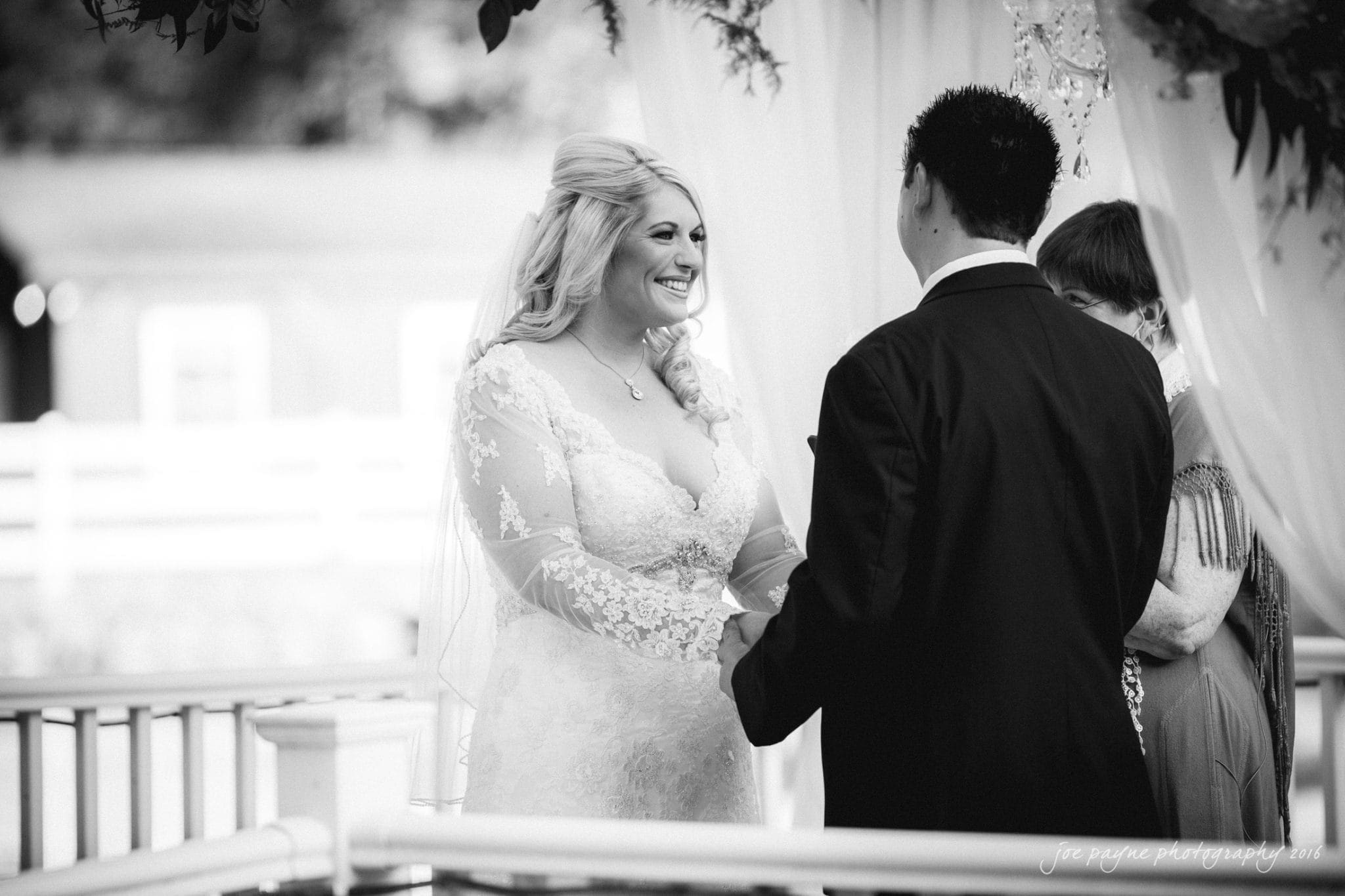 shady wagon farm wedding photography - danielle & josh A bride and groom stand facing each other, holding hands and smiling during an outdoor wedding ceremony. The bride is wearing a lace dress, and the groom is in a suit. A third person, partially visible, stands nearby. The setting is bright and elegant.
