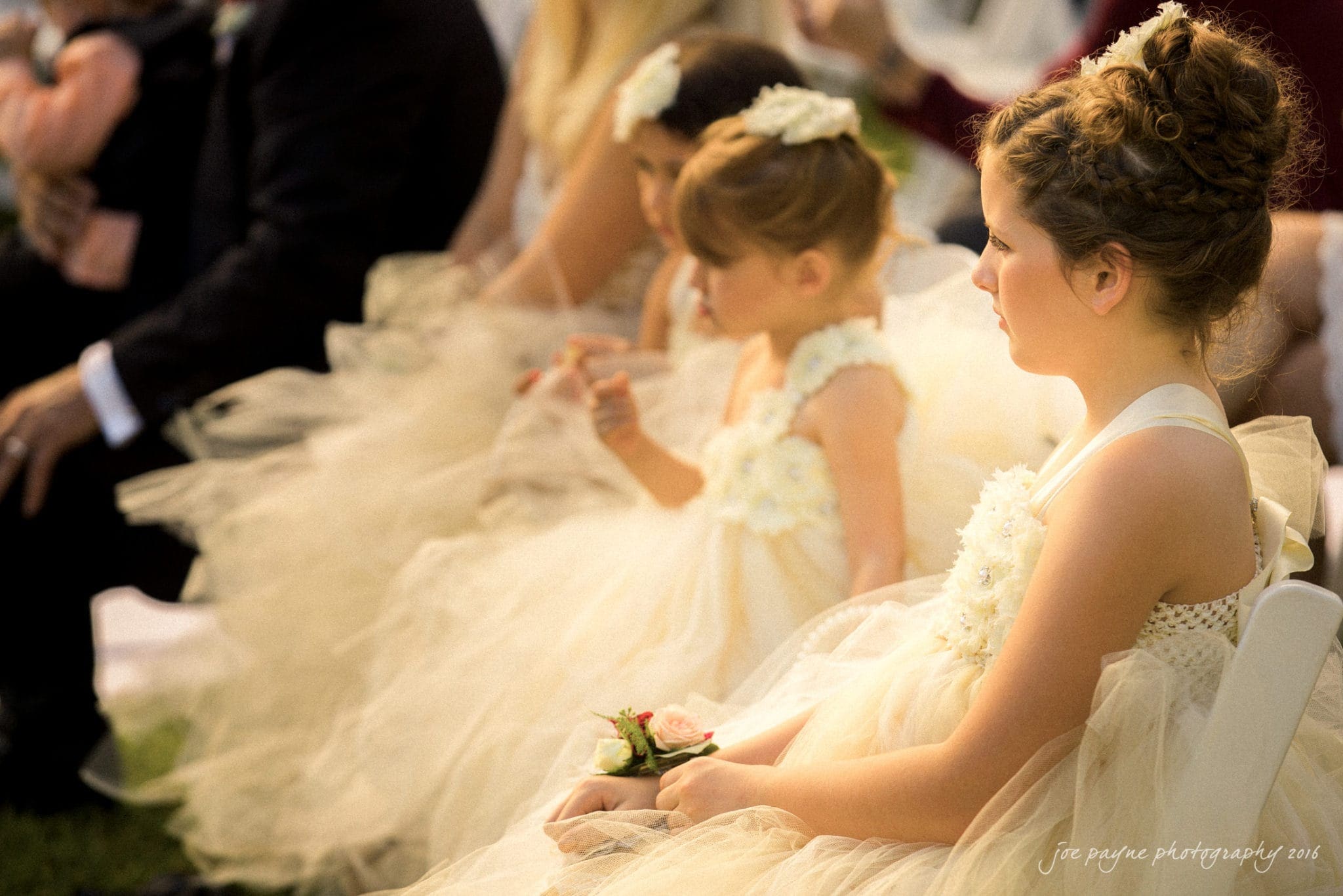 shady wagon farm wedding photography - danielle & josh A group of young girls in cream-colored dresses with floral details sit attentively at an outdoor event. One girl in the foreground holds a small bouquet with pink roses. Sunlight softly illuminates their surroundings.