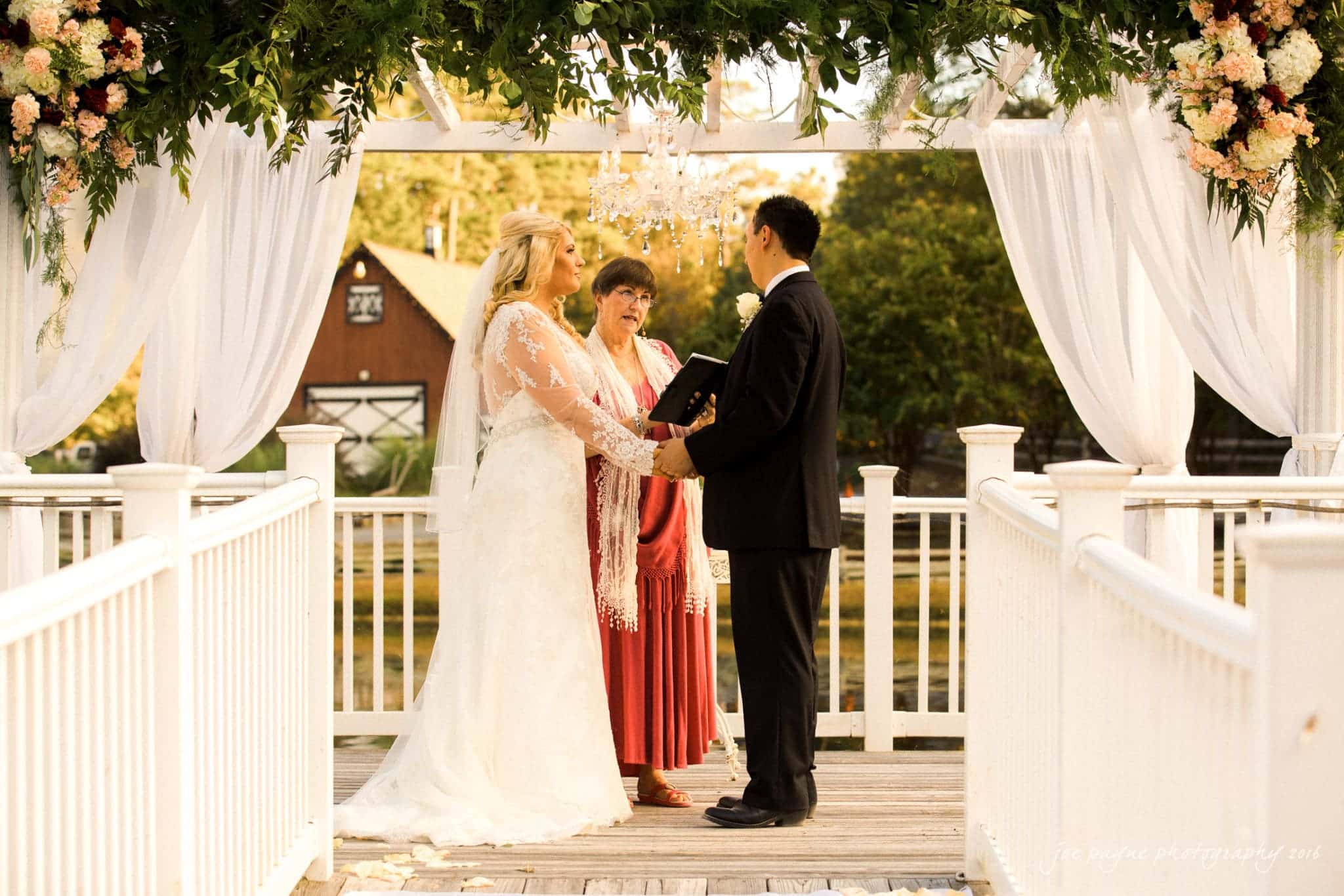 shady wagon farm wedding photography - danielle & josh A bride and groom are holding hands at an outdoor wedding ceremony under a floral and draped arch. A woman in a red dress stands between them, officiating. The setting is serene with a rustic barn and trees in the background.