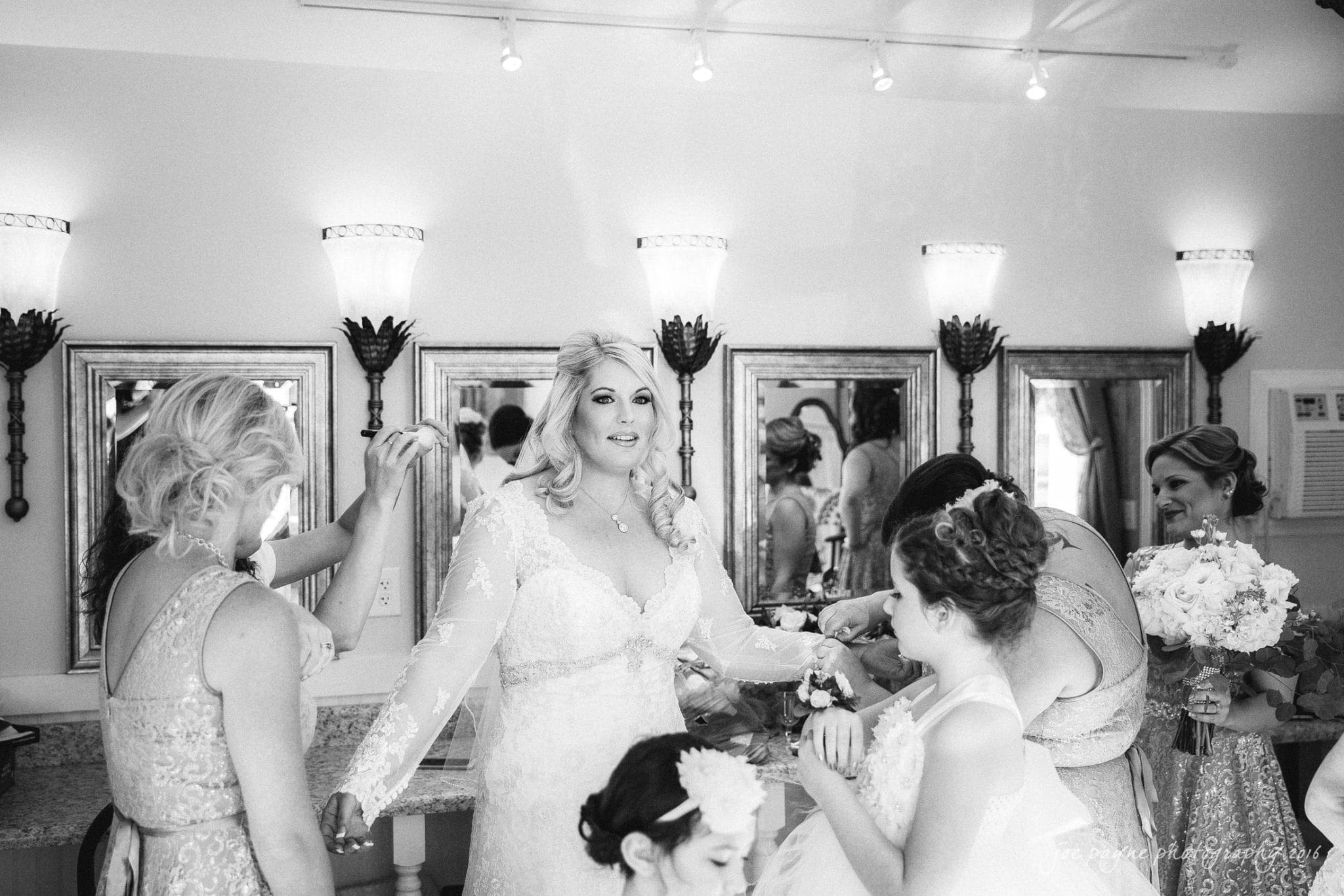 shady wagon farm wedding photography - danielle & josh A bride in a white gown and veil stands in a well-lit dressing room with chandeliers. She is surrounded by bridesmaids and children in matching dresses, helping her prepare. Smiling, she looks composed and ready for the ceremony.