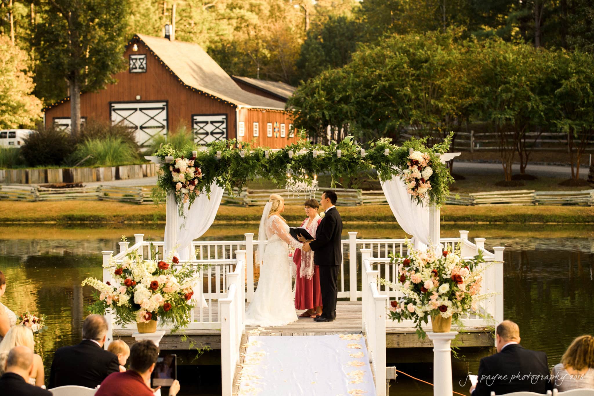 shady wagon farm wedding photography - danielle & josh A couple is getting married on a small white pier decorated with floral arrangements over a lake. Guests sit on both sides, and a rustic barn is in the background surrounded by trees.