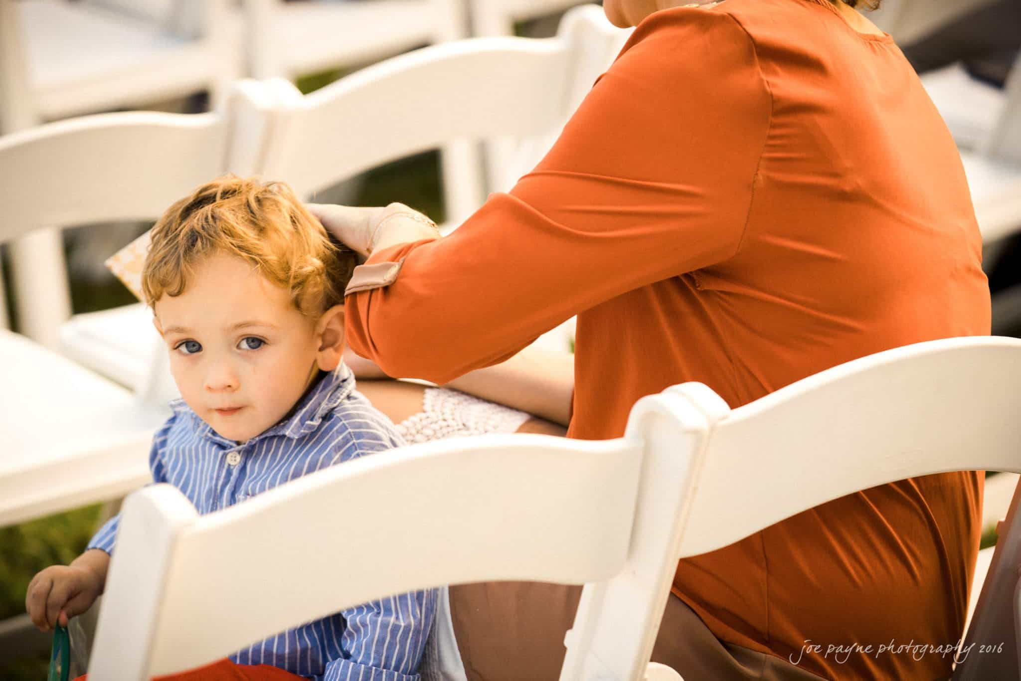 shady wagon farm wedding photography - danielle & josh A young child with curly hair and a blue striped shirt sits on a white chair, looking at the camera. An adult next to him, wearing an orange blouse, gently rests a hand on his head. Theyre outdoors with sunlight shining.