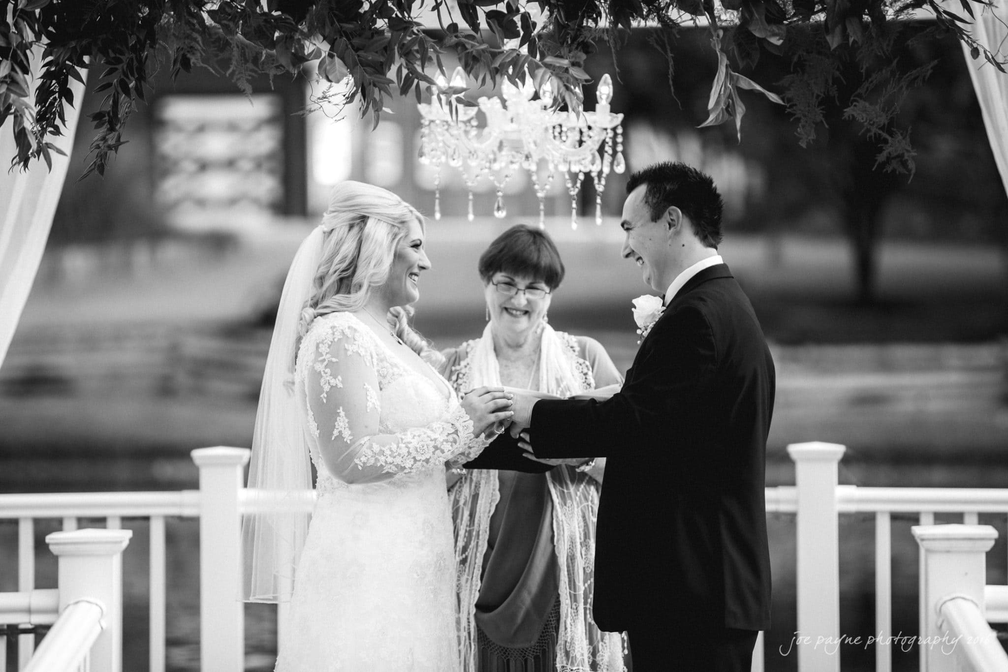 shady wagon farm wedding photography - danielle & josh A bride and groom stand facing each other, holding hands under a decorated gazebo. The officiant, smiling, stands between them. The setting is elegant, with a chandelier overhead and greenery in the background.