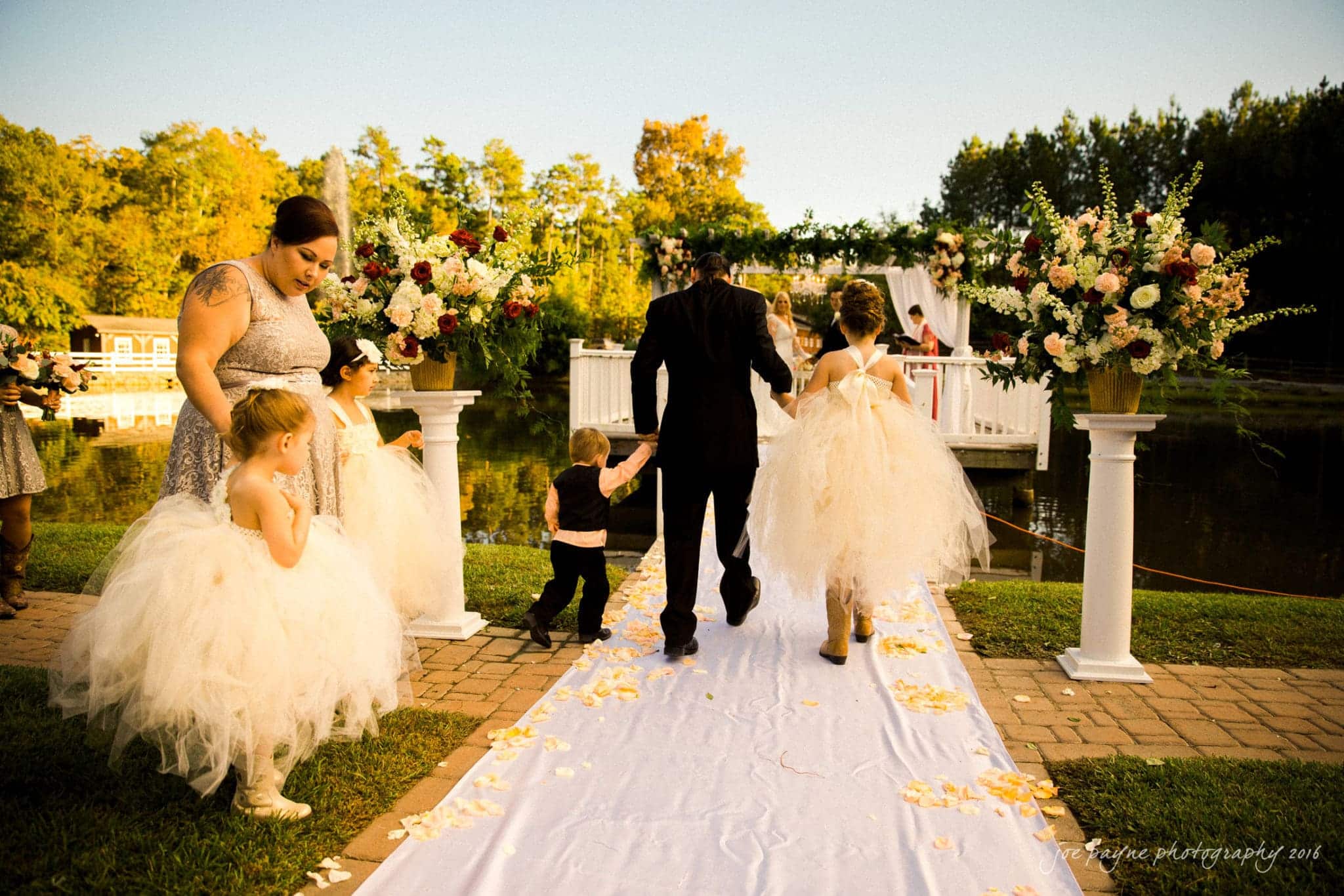 shady wagon farm wedding photography - danielle & josh A wedding party walks down an outdoor aisle by a lake. The flower girls wear white tulle dresses, and the ring bearer is in a black outfit. The path is lined with rose petals, and large floral arrangements adorn the scene.