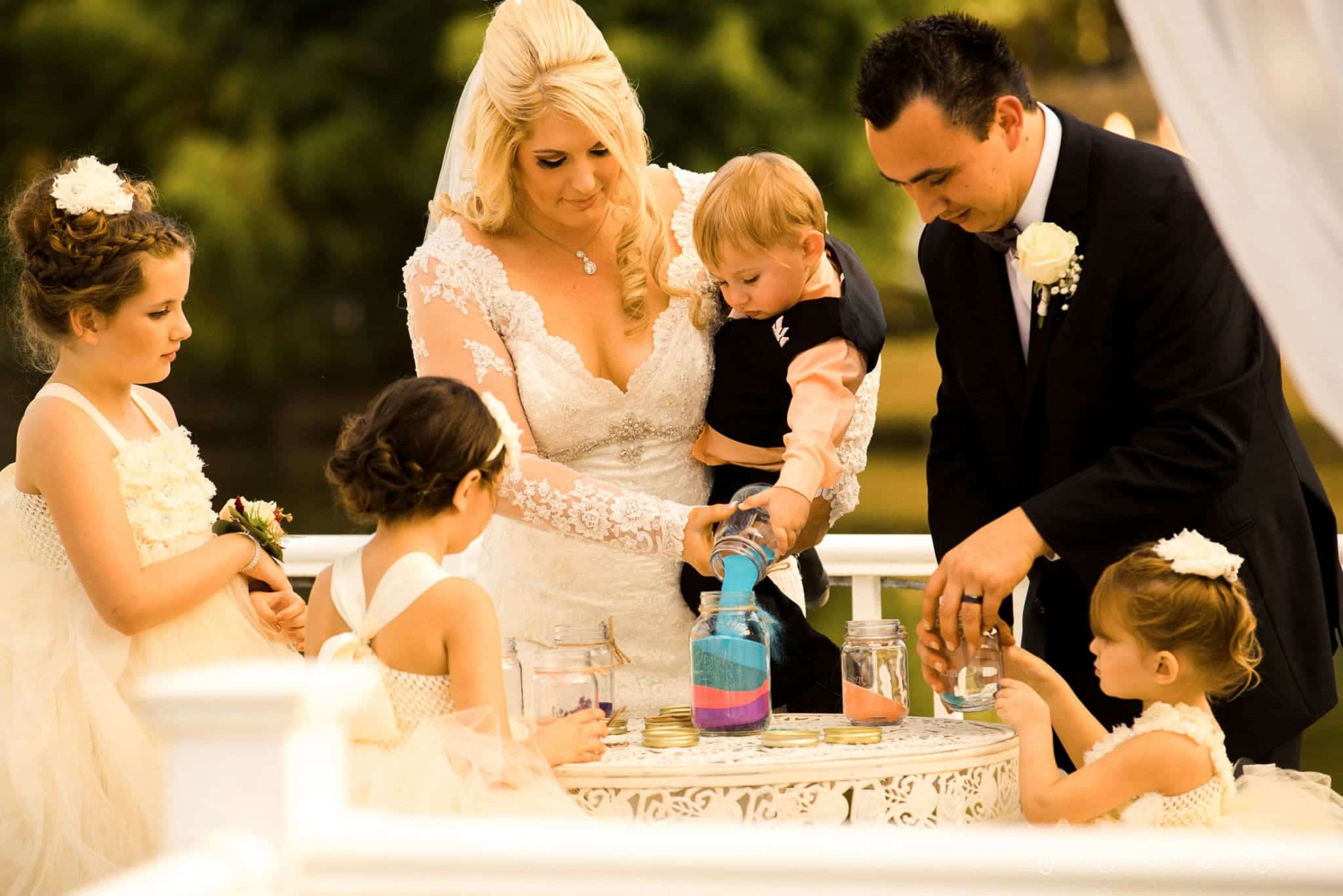 shady wagon farm wedding photography - danielle & josh A family participates in a sand ceremony during a wedding. The bride holds a baby, and the groom pours sand. Two young girls in white dresses watch, and another girl stands nearby, all gathered around a table with jars of colored sand.