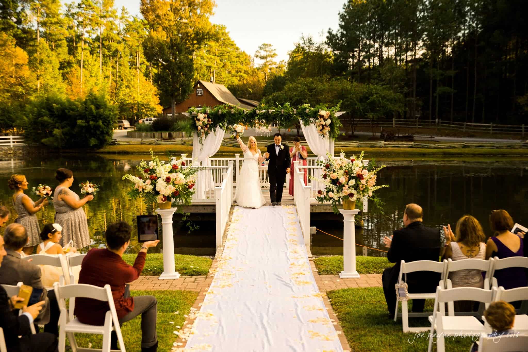 shady wagon farm wedding photography - danielle & josh A bride and groom walk down an aisle on a white bridge adorned with flowers, surrounded by seated guests. The setting is outdoors, near a pond, with trees and a rustic building in the background under a clear sky.
