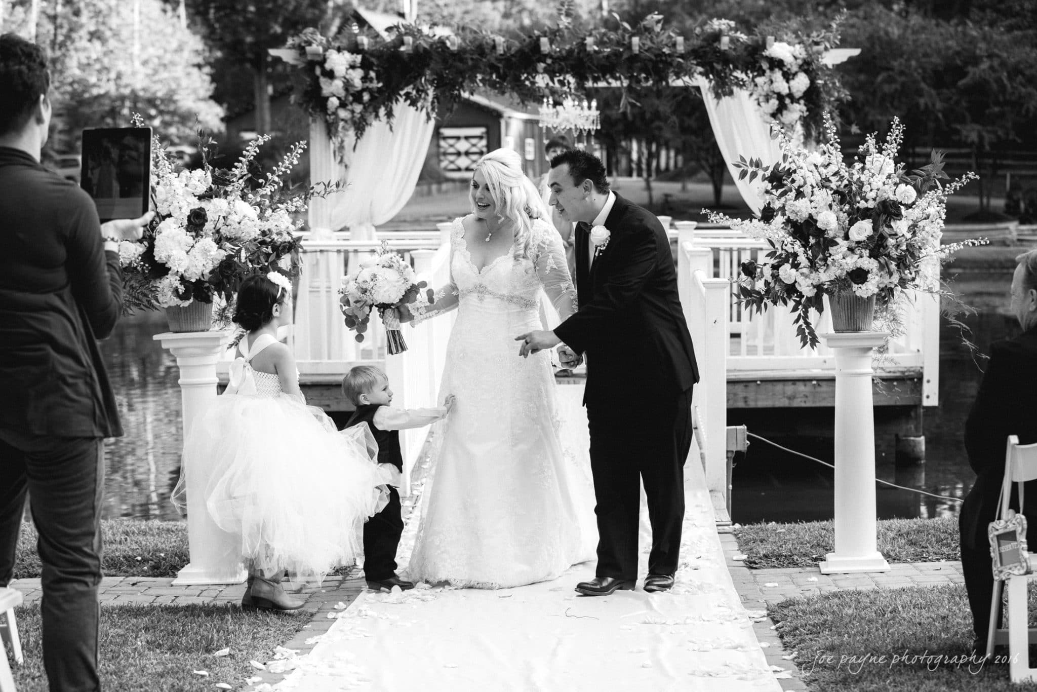 shady wagon farm wedding photography - danielle & josh A bride and groom stand smiling under a floral arch at an outdoor wedding, holding hands with two young children. The scene is captured in black and white, with trees and water in the background.