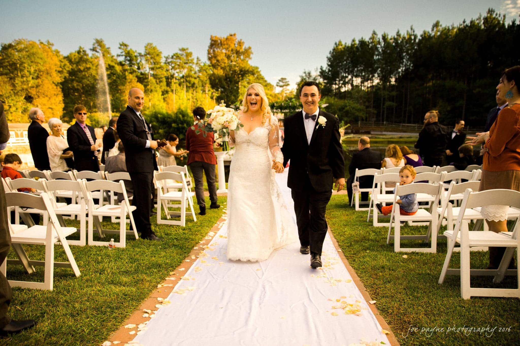 shady wagon farm wedding photography - danielle & josh A bride and groom joyfully walk down an outdoor aisle lined with white chairs and flower petals. Guests are seated on both sides, some clapping. Theres a green lawn and trees in the background under a clear sky.