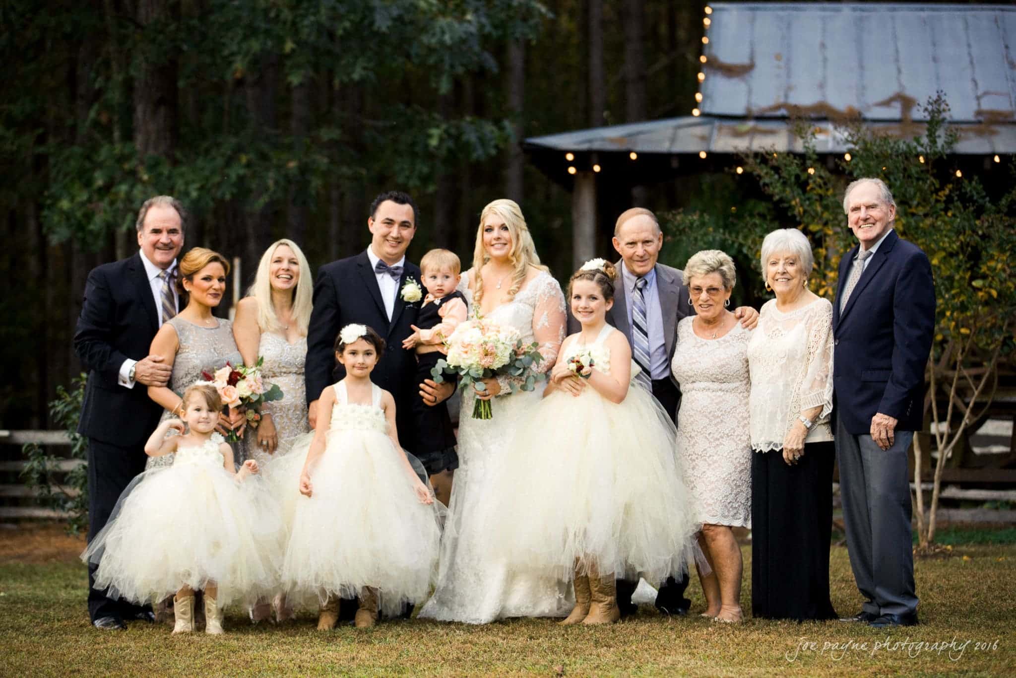 shady wagon farm wedding photography - danielle & josh A large wedding group stands outdoors. The bride and groom are in the center holding a child. Bridesmaids in white dresses stand to the left, and family members in formal attire are positioned on either side. A rustic building is in the background.