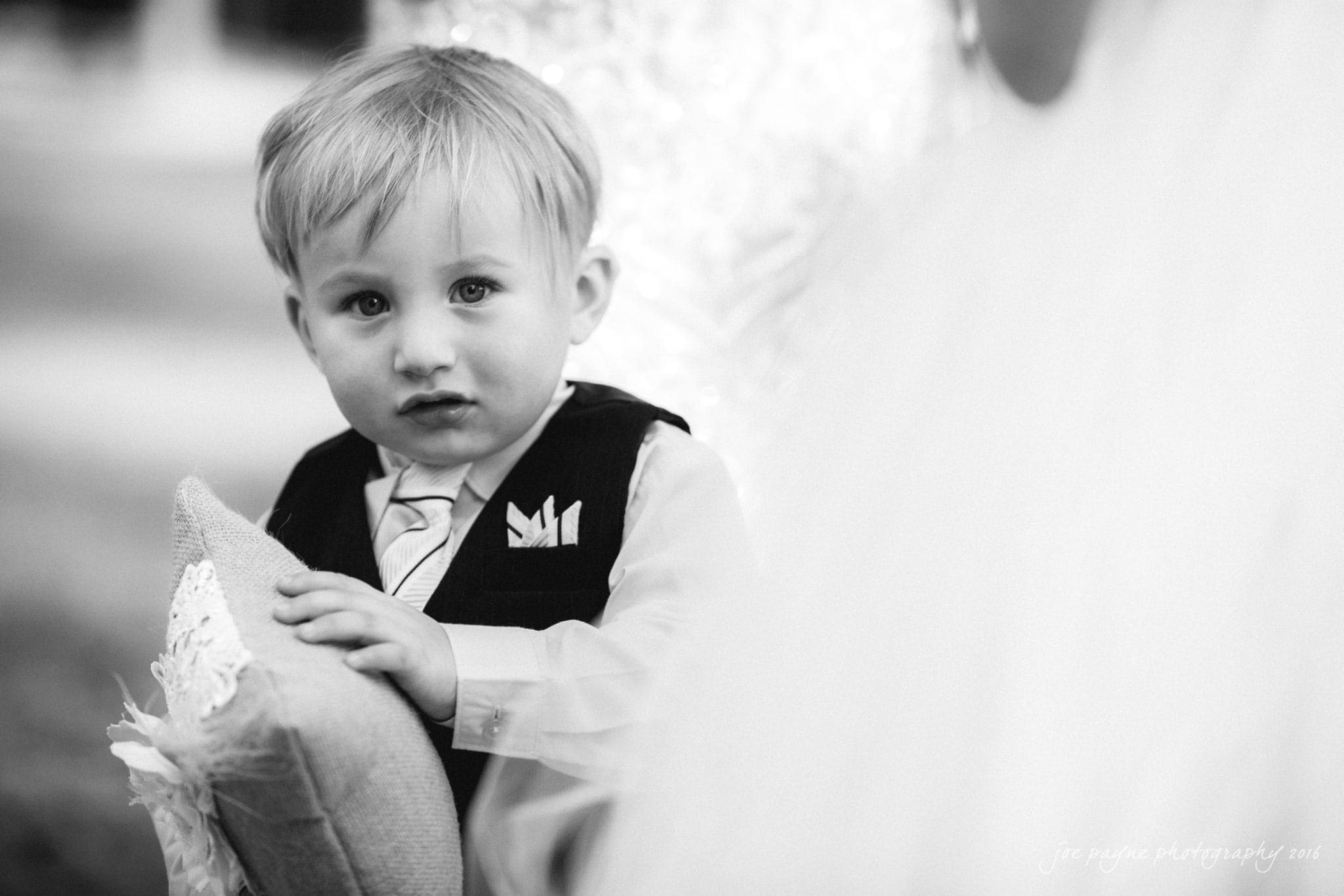 shady wagon farm wedding photography - danielle & josh A young boy in formal attire holds a decorative pillow, looking intently towards the camera. The background is softly blurred, suggesting an outdoor setting. The photo is in black and white, highlighting the boys expression.