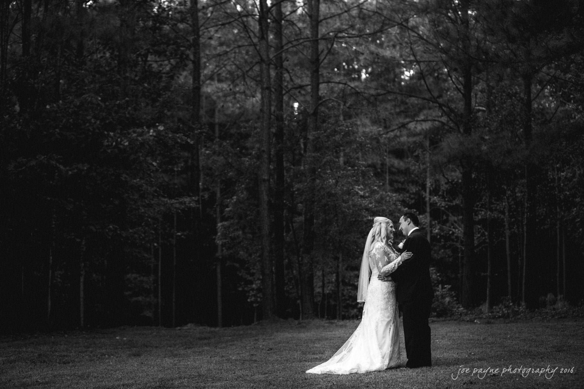 shady wagon farm wedding photography - danielle & josh A bride and groom embrace on a grassy field surrounded by tall trees. The image is in black and white, adding a classic and romantic atmosphere to the scene.