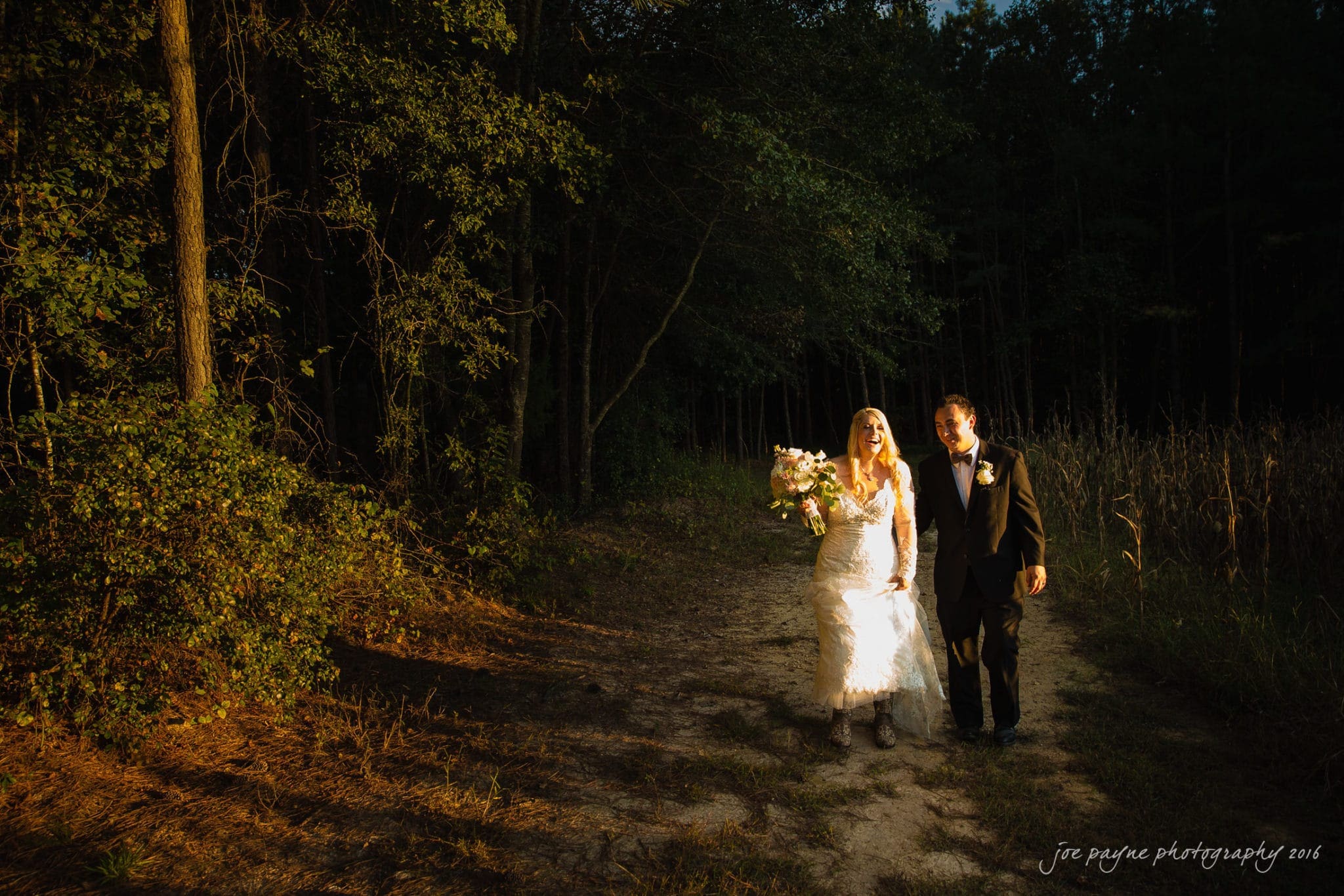 shady wagon farm wedding photography - danielle & josh A bride and groom walk hand-in-hand along a sunlit forest path. The bride wears a white lace gown and holds a bouquet of flowers, while the groom wears a dark suit. The surrounding trees are illuminated by warm evening light.
