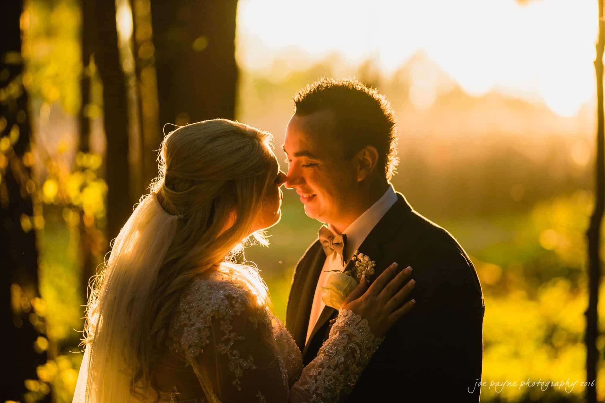 shady wagon farm wedding photography - danielle & josh A bride and groom stand close together in a sunlit forest. The groom wears a dark suit, and the bride wears a lace wedding dress. They are gazing at each other, surrounded by warm, golden light filtering through the trees.