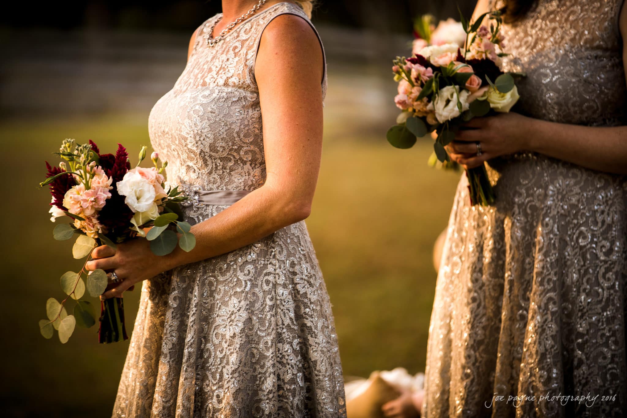 shady wagon farm wedding photography - danielle & josh Two bridesmaids in lace dresses hold bouquets of pink and white flowers with greenery. The warm, golden light enhances the elegance of the scene. A wooden fence and blurred background suggest an outdoor setting.
