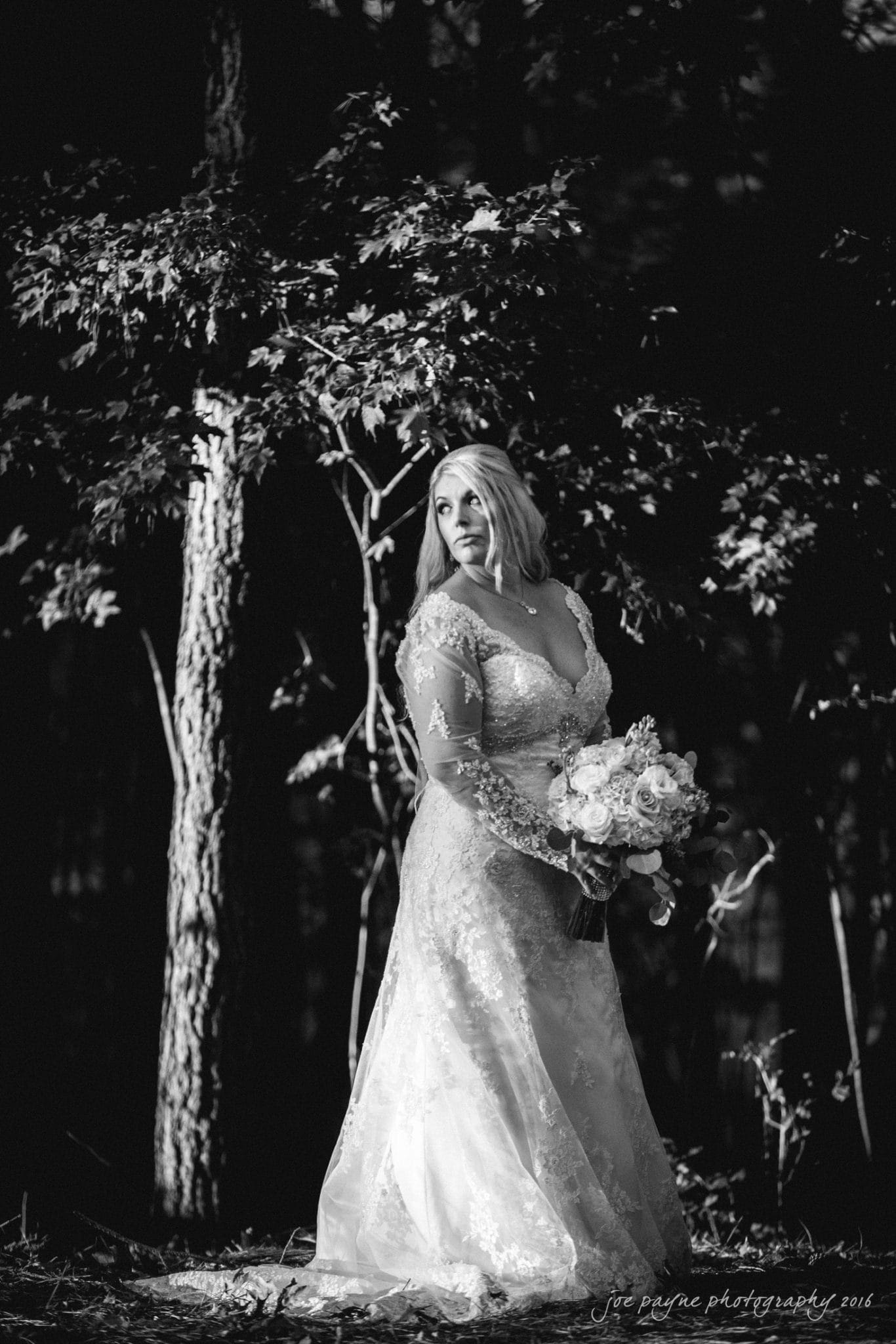 shady wagon farm wedding photography - danielle & josh A bride in an elegant lace wedding dress stands in a forest, holding a bouquet of flowers. Dappled sunlight filters through the trees, illuminating her and creating a serene, romantic atmosphere.