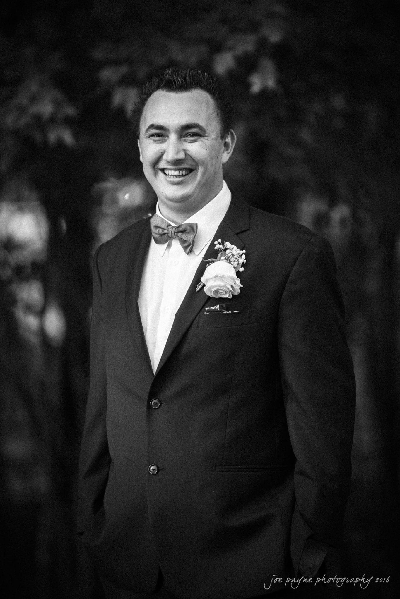 shady wagon farm wedding photography - danielle & josh A man in a suit and bow tie smiles warmly in a black and white photo. He wears a boutonnière on his lapel. The background is softly blurred with hints of foliage.